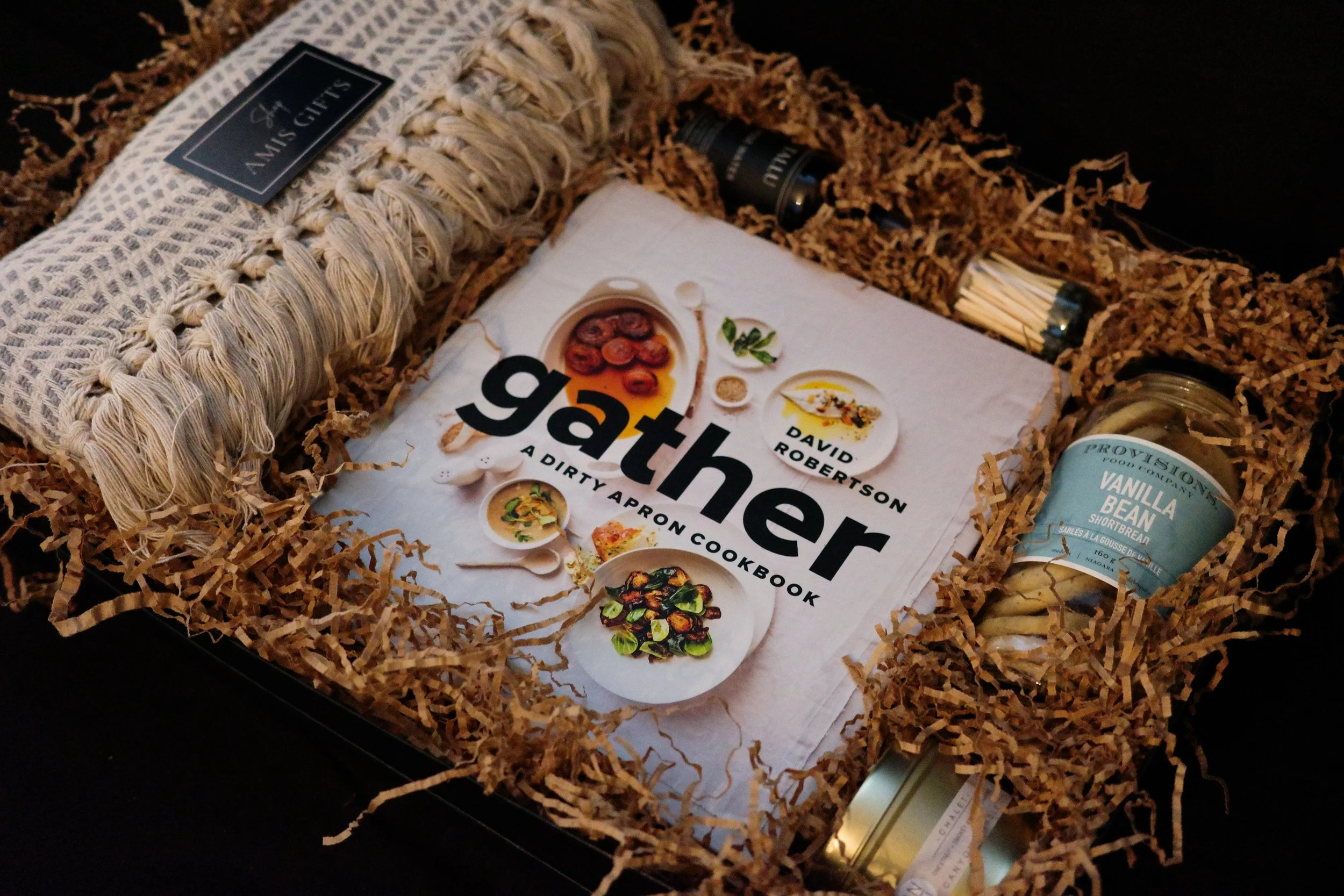 Gift box filled with shredded paper, containing a cookbook titled 'GATHER: A DIRTY APRON COOKBOOK' by David Robertson, a bottle of Provigo vanilla bean shortbread, a small jar, and a black container.