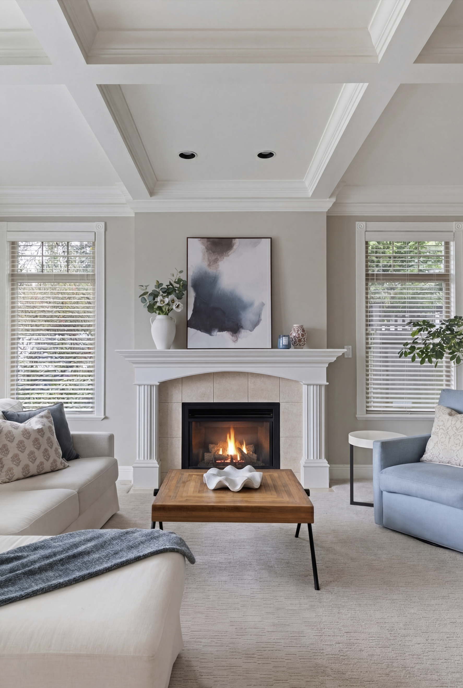 Living room with a fireplace, beige couch with patterned pillows, and blue armchair, wooden coffee table with white decor, large abstract painting above fireplace, two windows with white blinds, and decorative vases and plant on shelves.