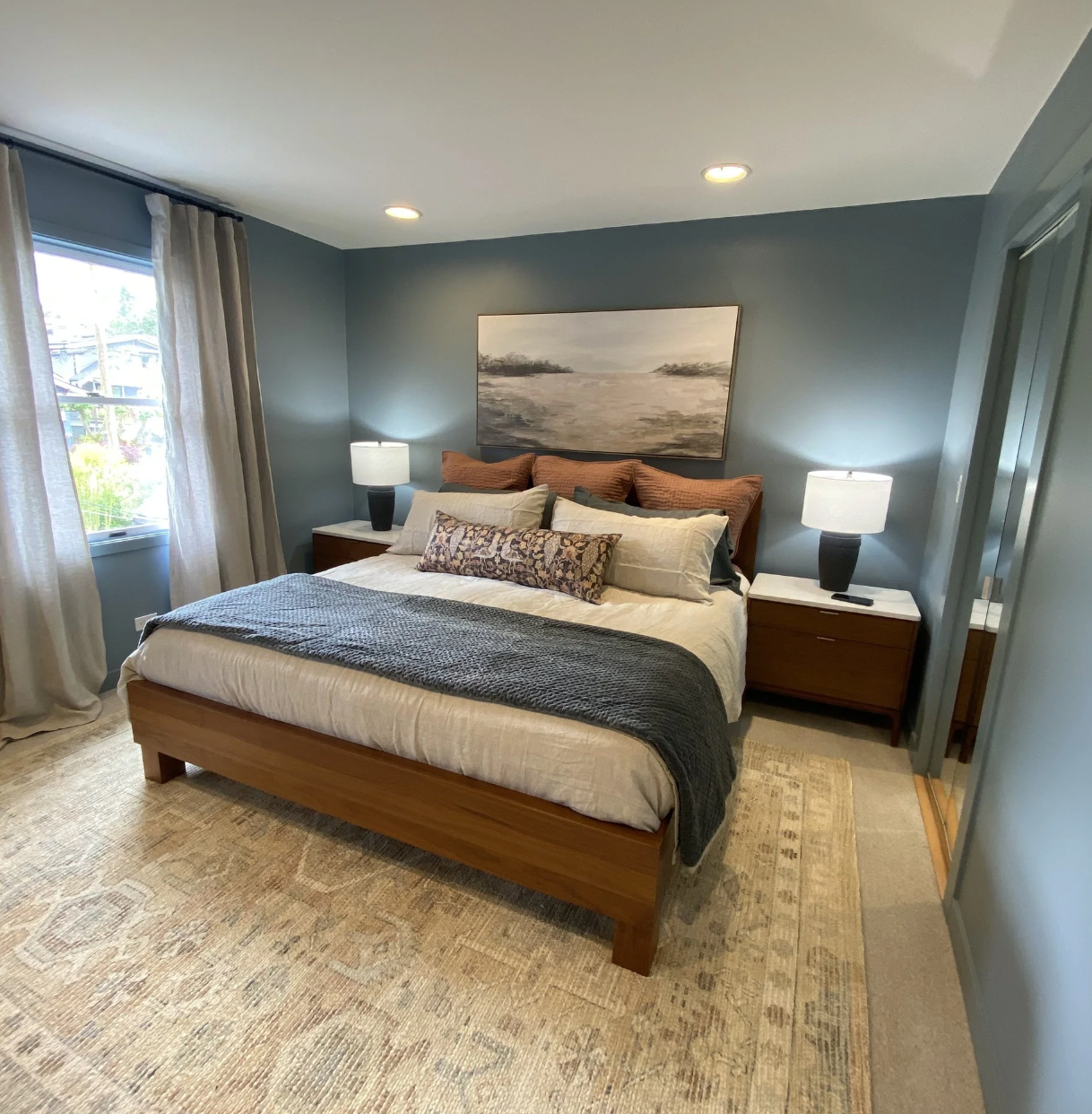 A bedroom with a wooden bed frame, gray bedding, multiple decorative pillows, two nightstands with lamps, a window with curtains, a painting above the bed, and a beige rug.