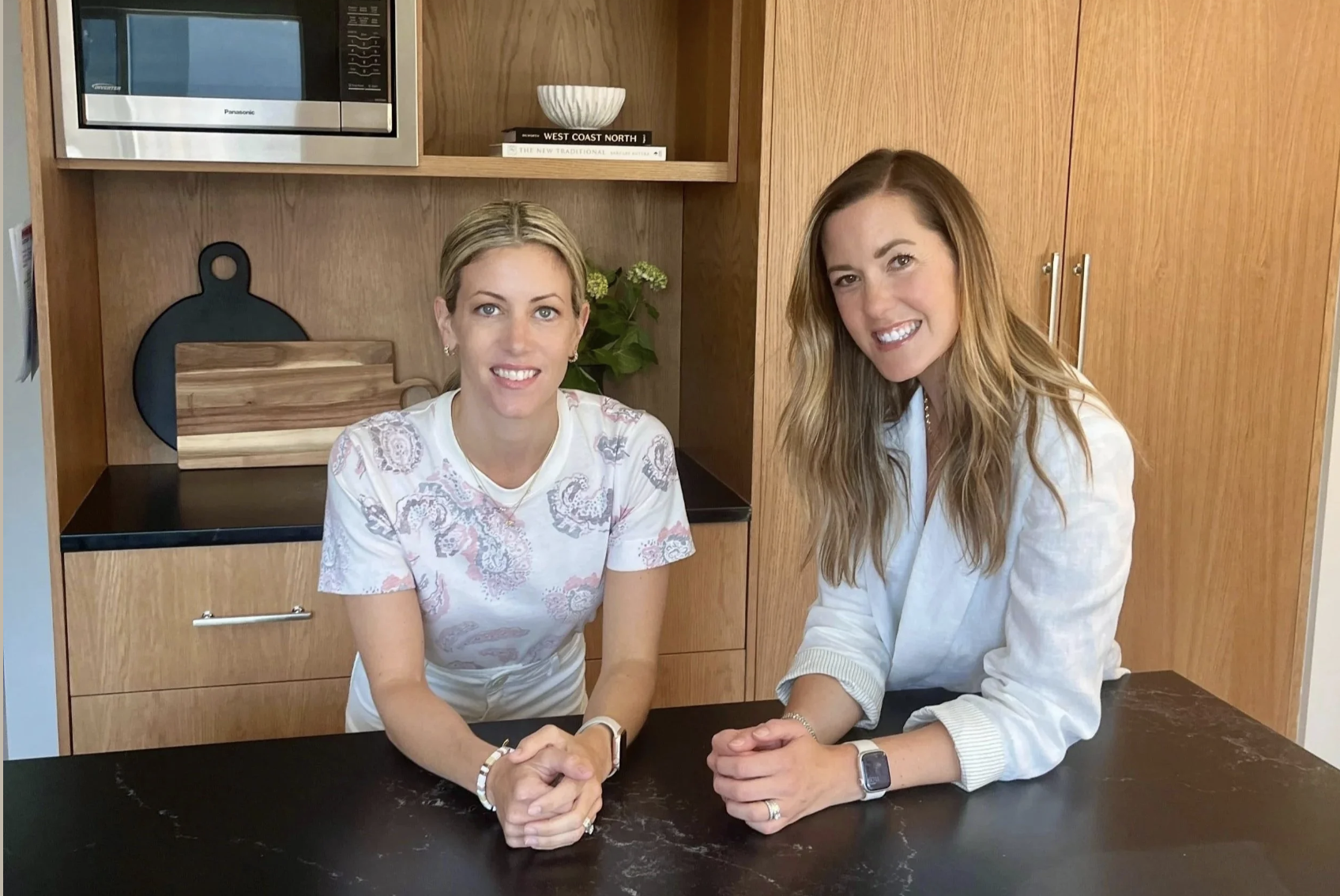 Two women sitting at a dark counter or table in a kitchen, smiling at the camera. One has blonde hair and is wearing a white patterned shirt, and the other has light brown hair and is wearing a white blazer.
