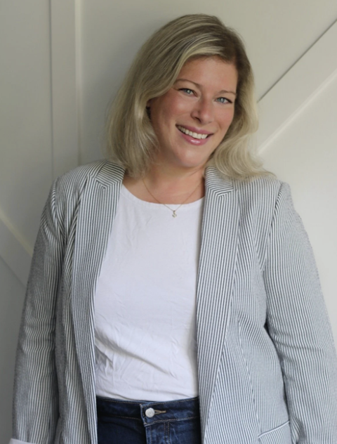 A woman with blonde hair smiling, wearing a white shirt and a gray striped blazer, standing in front of a neutral background.