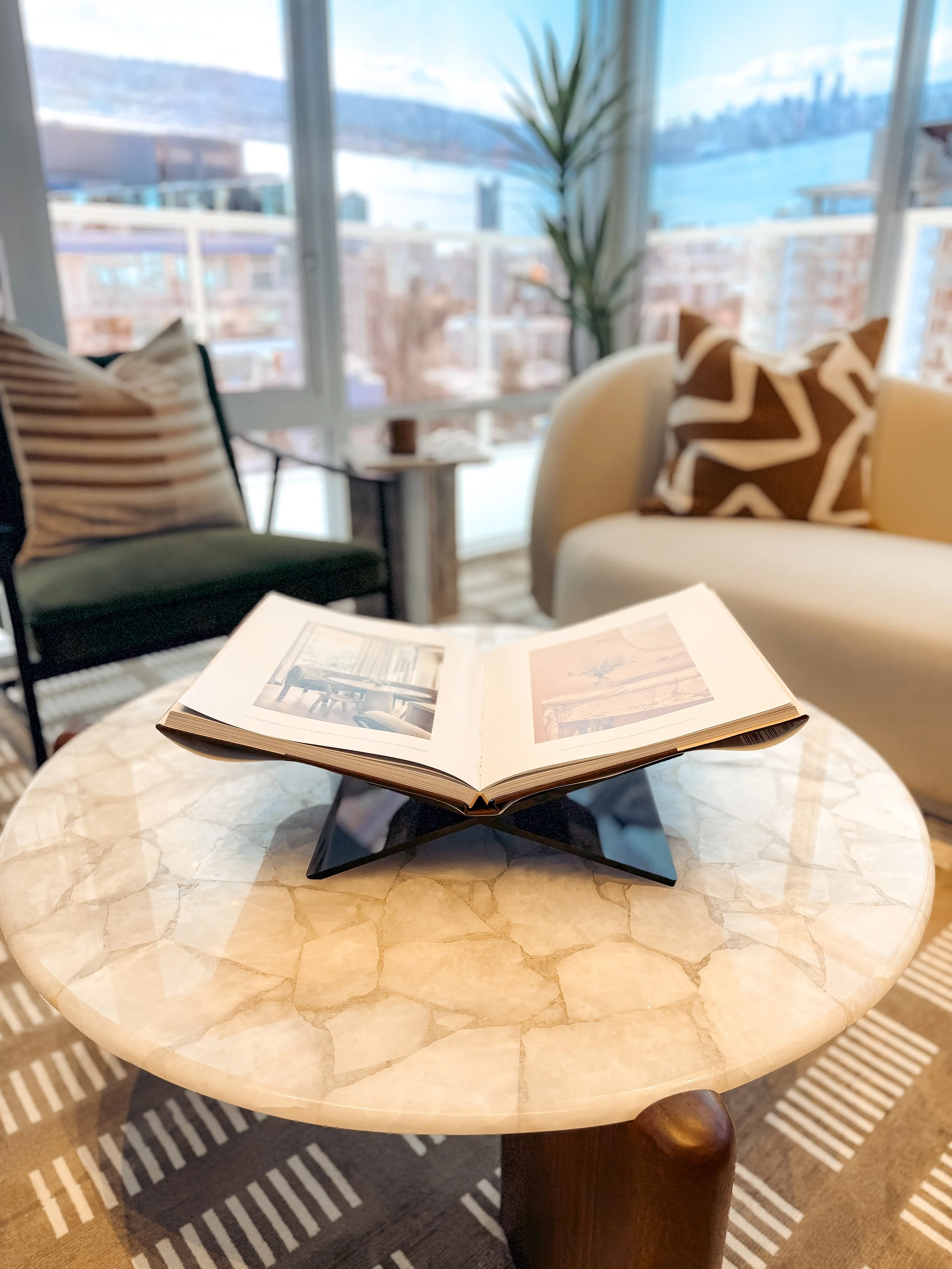 Open photo album placed on a decorative round marble table in a cozy living room with large windows, a chair with striped pillows, a yellow sofa with zigzag-patterned cushions, and a tall plant in the background overlooking a cityscape and mountains.