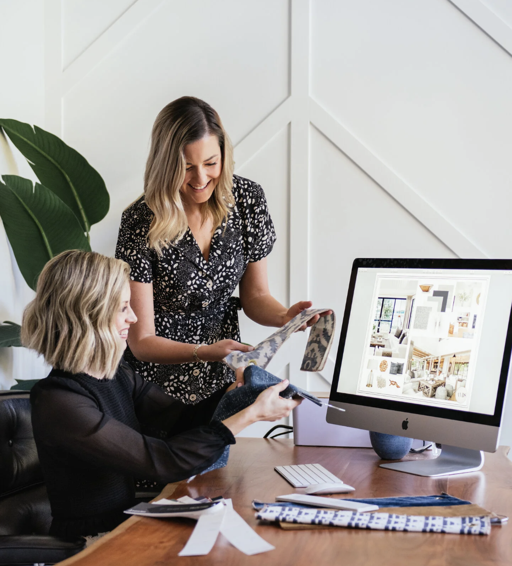 Two women discussing fabric samples in a design office, with a computer displaying interior design images.