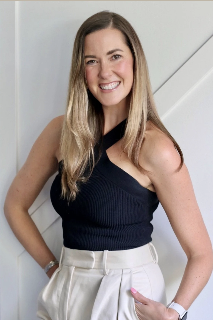 A woman with long blonde hair, wearing a black sleeveless top and white high-waisted pants, smiling and posing with one hand in her pocket.