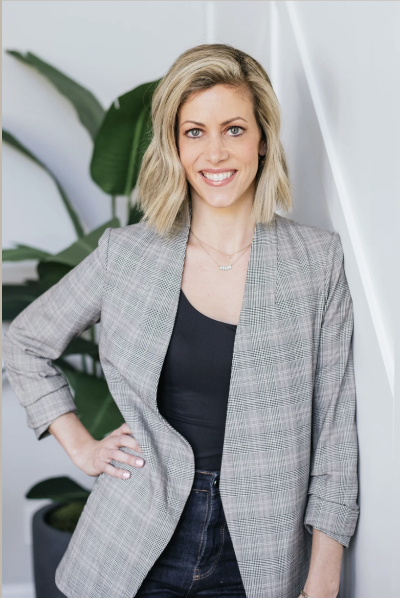 A woman with blonde hair smiling, wearing a gray plaid blazer and a black top, standing indoor near a large green plant and a white wall.