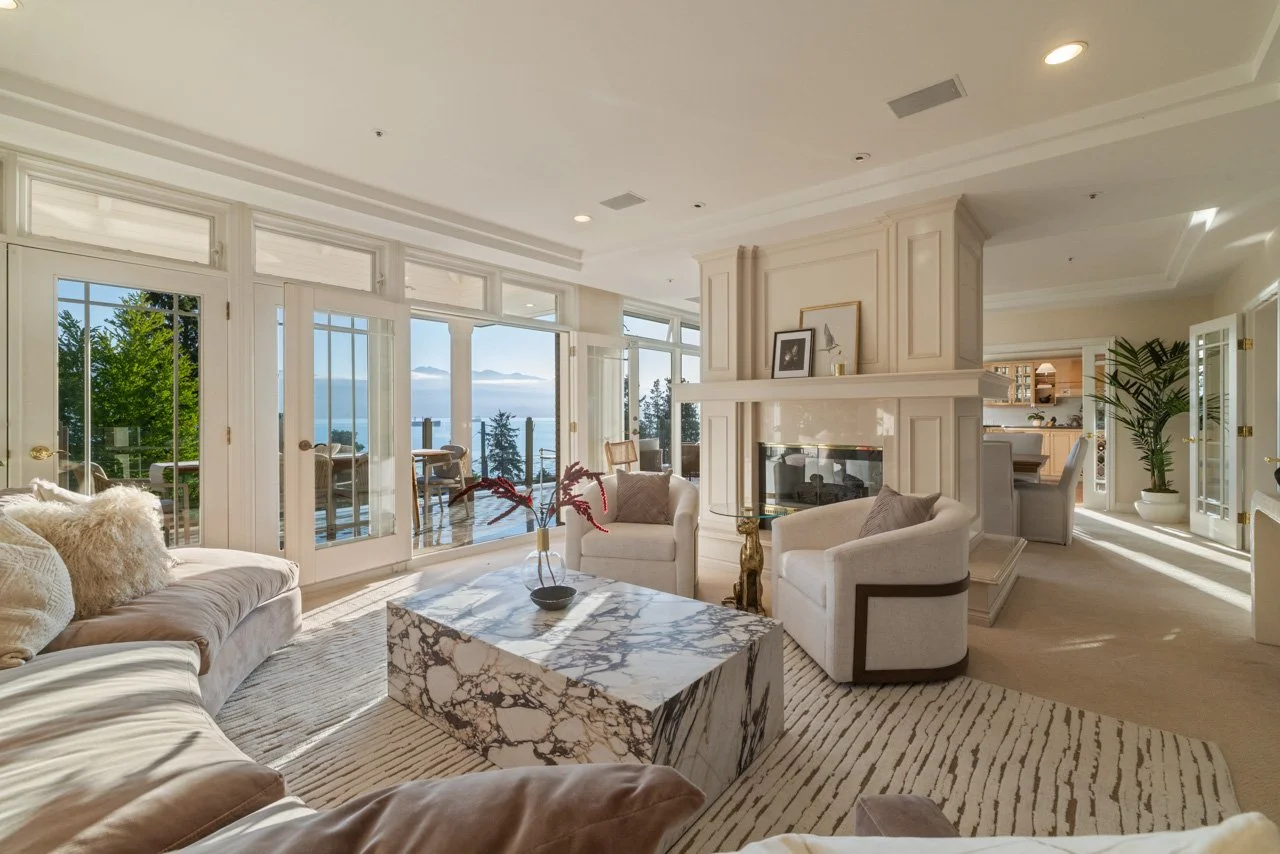 Bright living room with large windows overlooking a forest and lake, beige and white furniture, marble coffee table, and a fireplace.