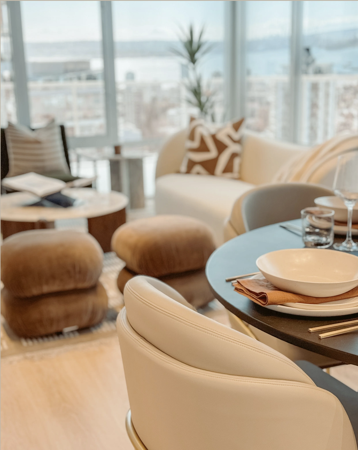 A cozy living room and dining area with large windows, beige and brown furniture, a round dining table set with plates, glasses, and utensils, and a view of buildings and water outside.