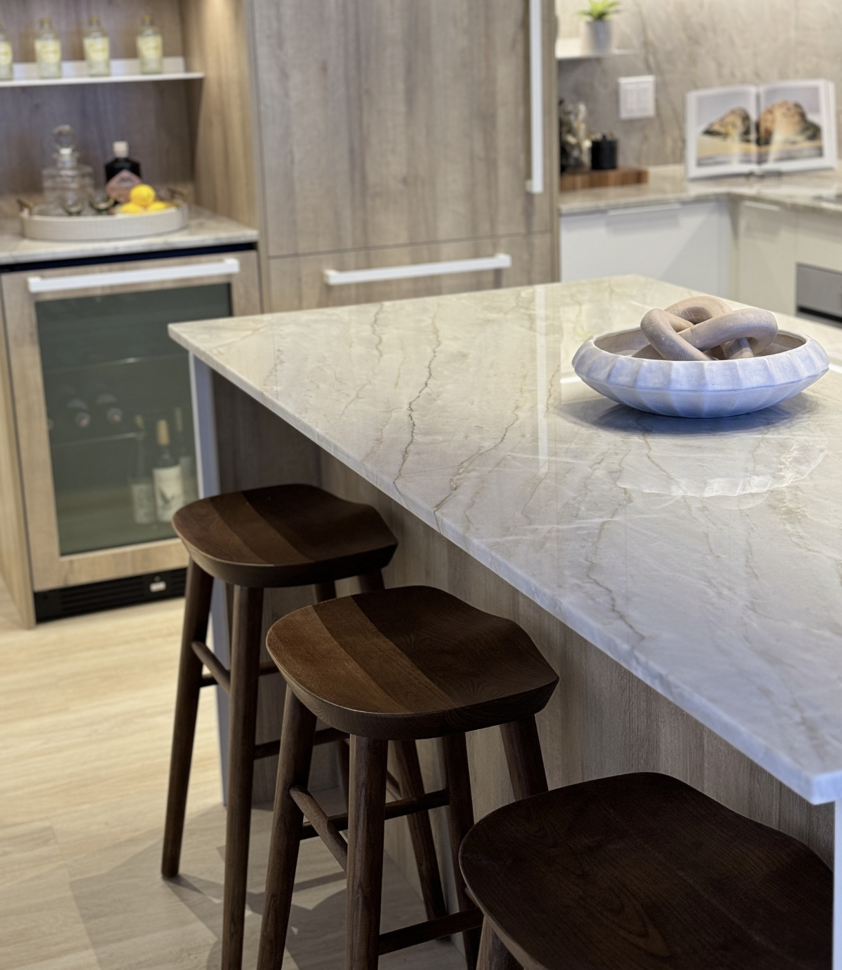 Modern kitchen with a marble island counter, wooden bar stools, a bowl with rope, and a beverage fridge, with additional kitchen appliances and decor in the background.