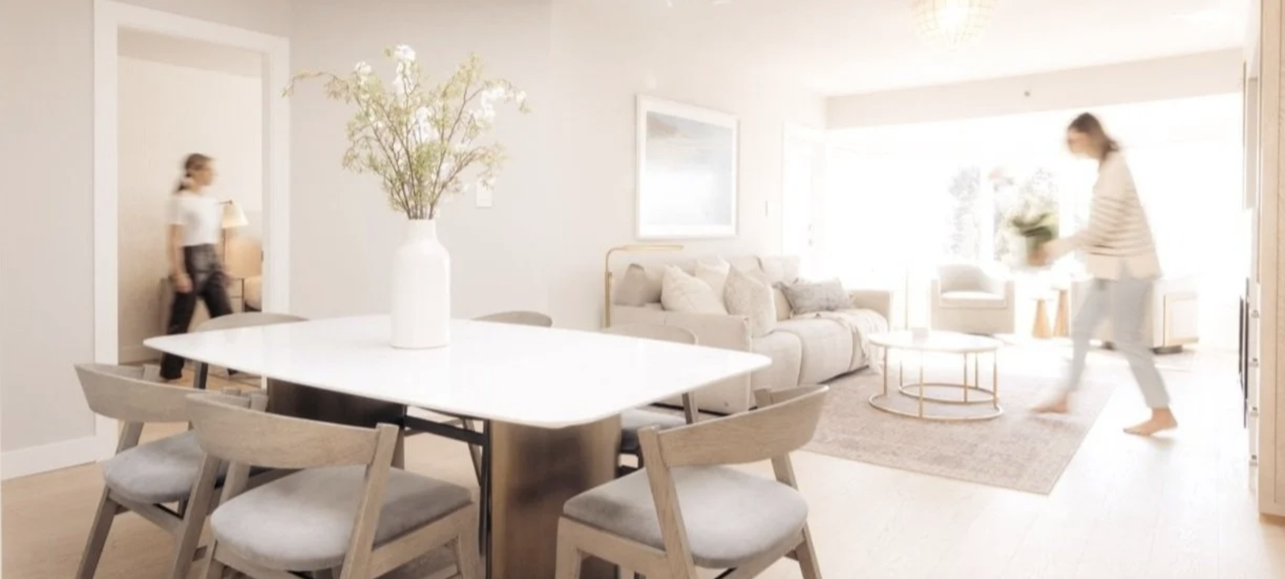 Bright living room with a beige sofa, a round coffee table, and a large window letting in natural light. A woman is near the entrance, and a young girl is walking away, both casually dressed. A white dining table with wooden chairs and a large white vase with flowers are also visible.