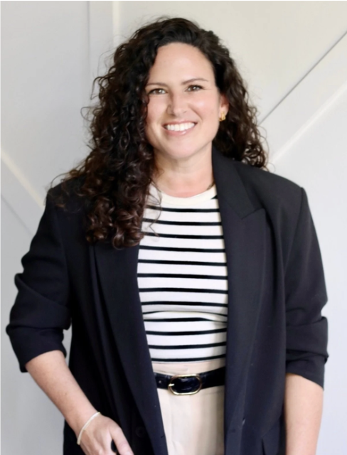 Woman with curly brown hair smiling, wearing a black blazer over a striped shirt, standing indoors.
