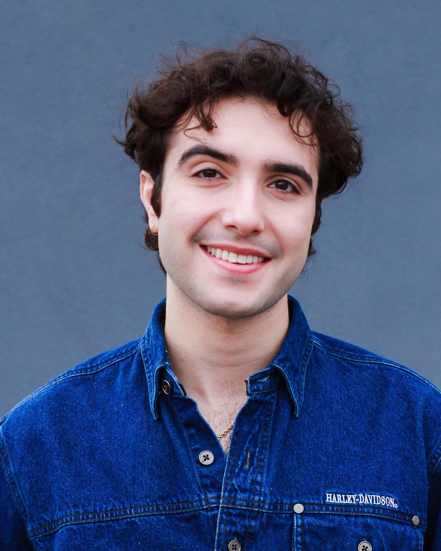 Young man with curly brown hair, fair skin, smiling, wearing a blue denim shirt with