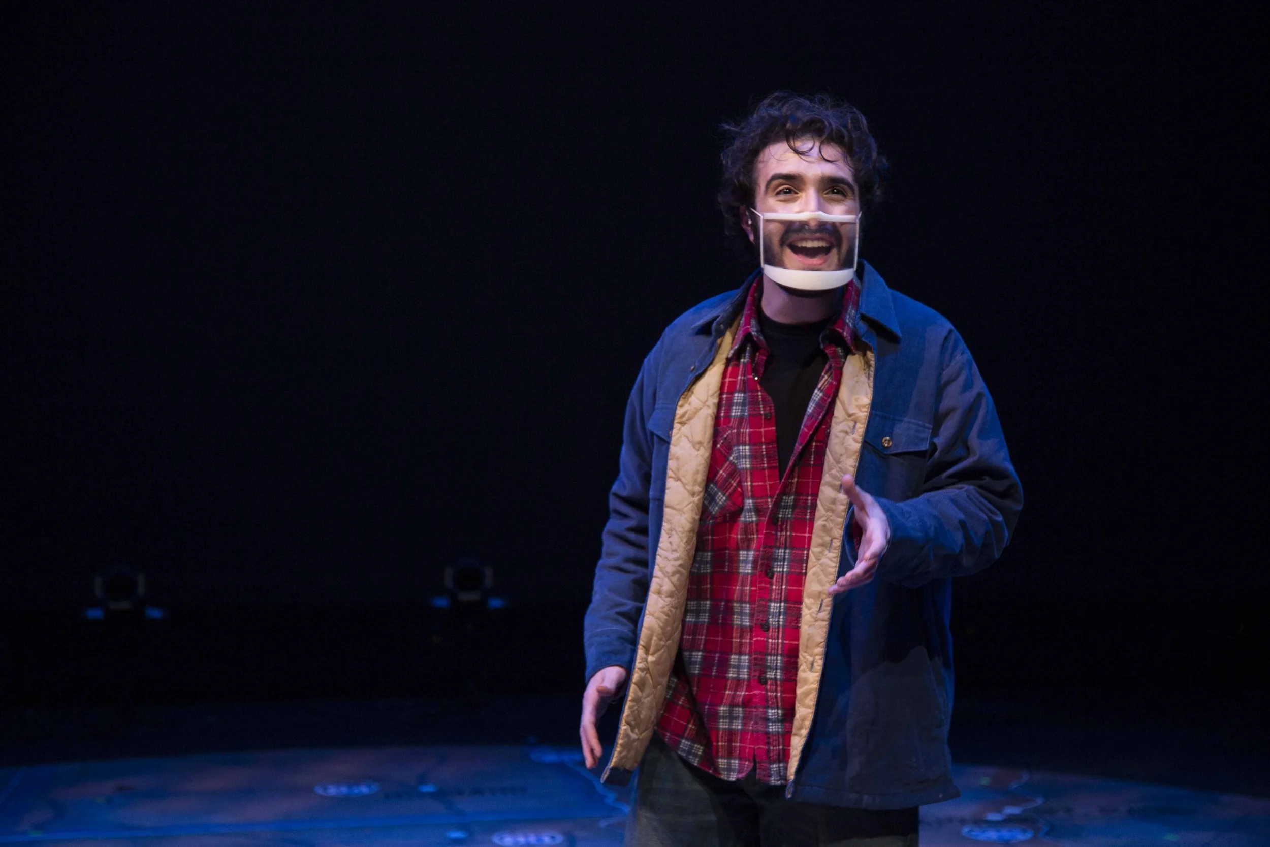A man on stage wearing a transparent face shield, a red plaid shirt, and a blue jacket, with a happy expression and gesturing with his right hand.