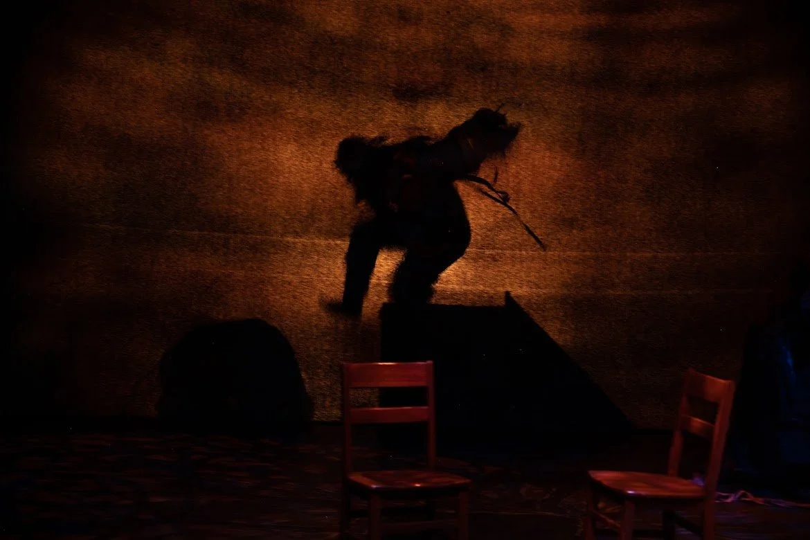 A silhouette of a person with a backpack and a stick, appearing to walk on a stage with orange lighting, with two chairs visible in the foreground.