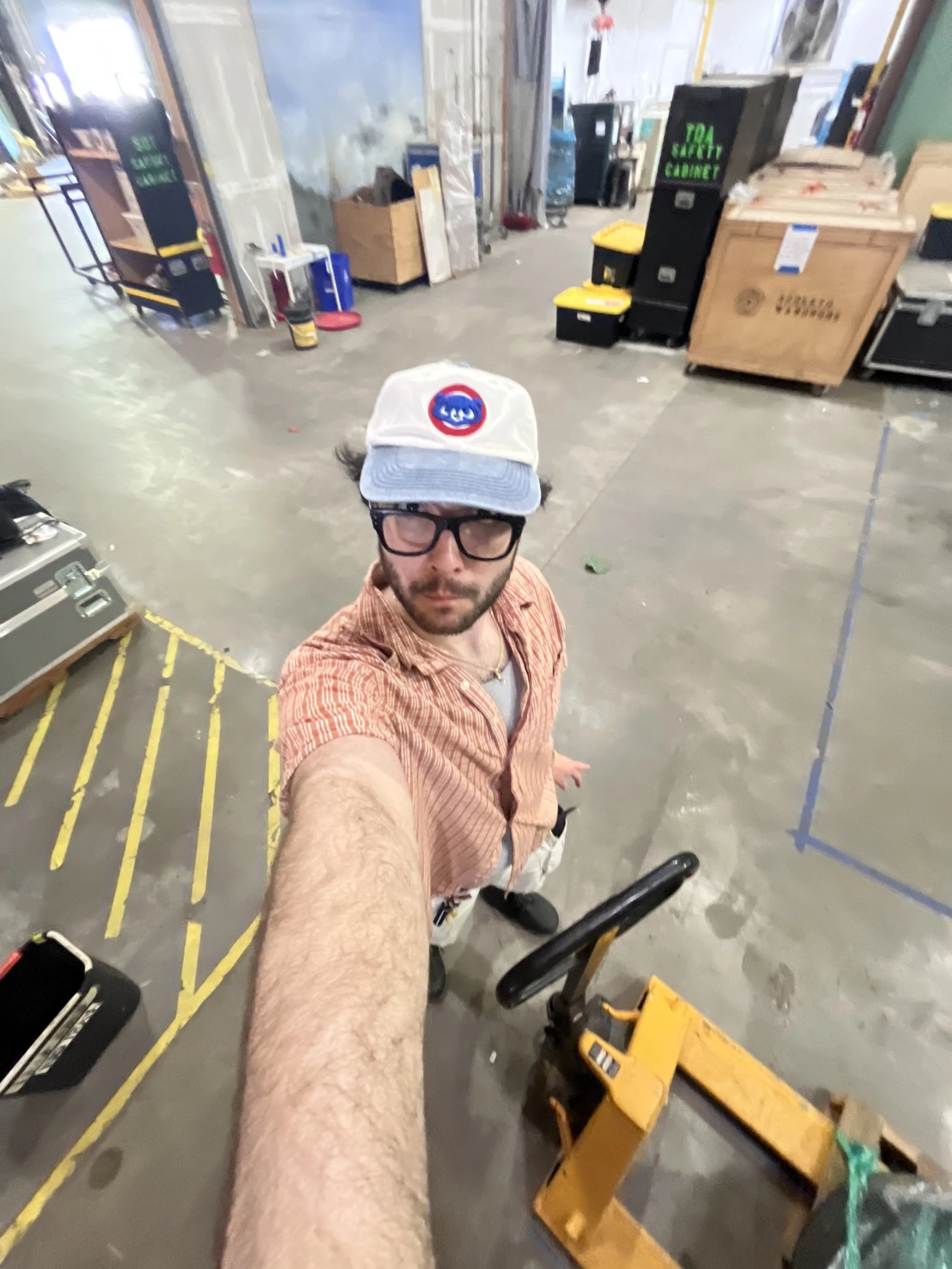 Selfie of a man wearing glasses, a white cap with a logo, and an orange striped shirt in an industrial or warehouse setting with boxes and equipment in the background.