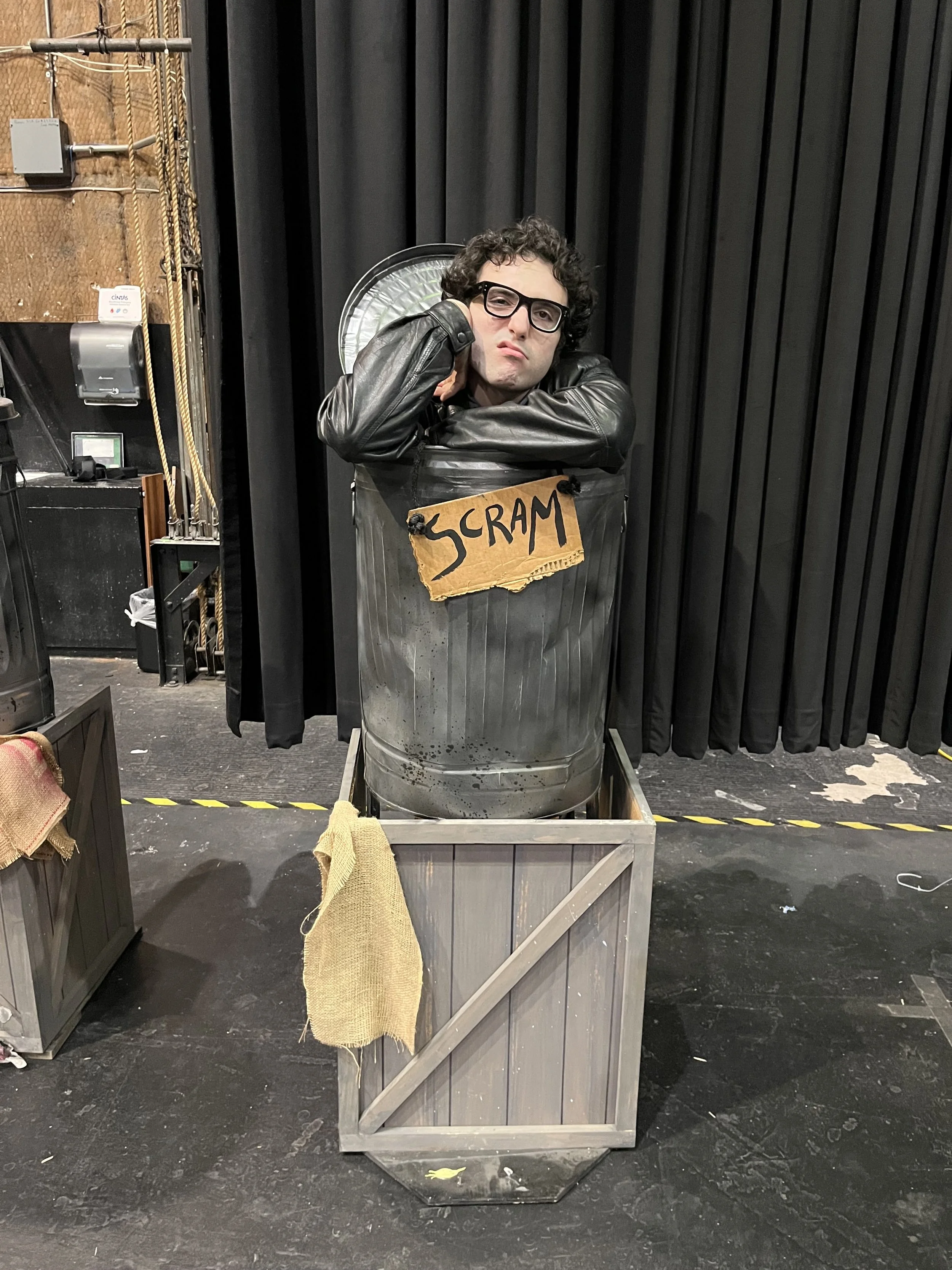 Person with curly hair and glasses leaning on a trash can with a 'SCRAM' sign, dressed as a character in a theatrical or stage setting.