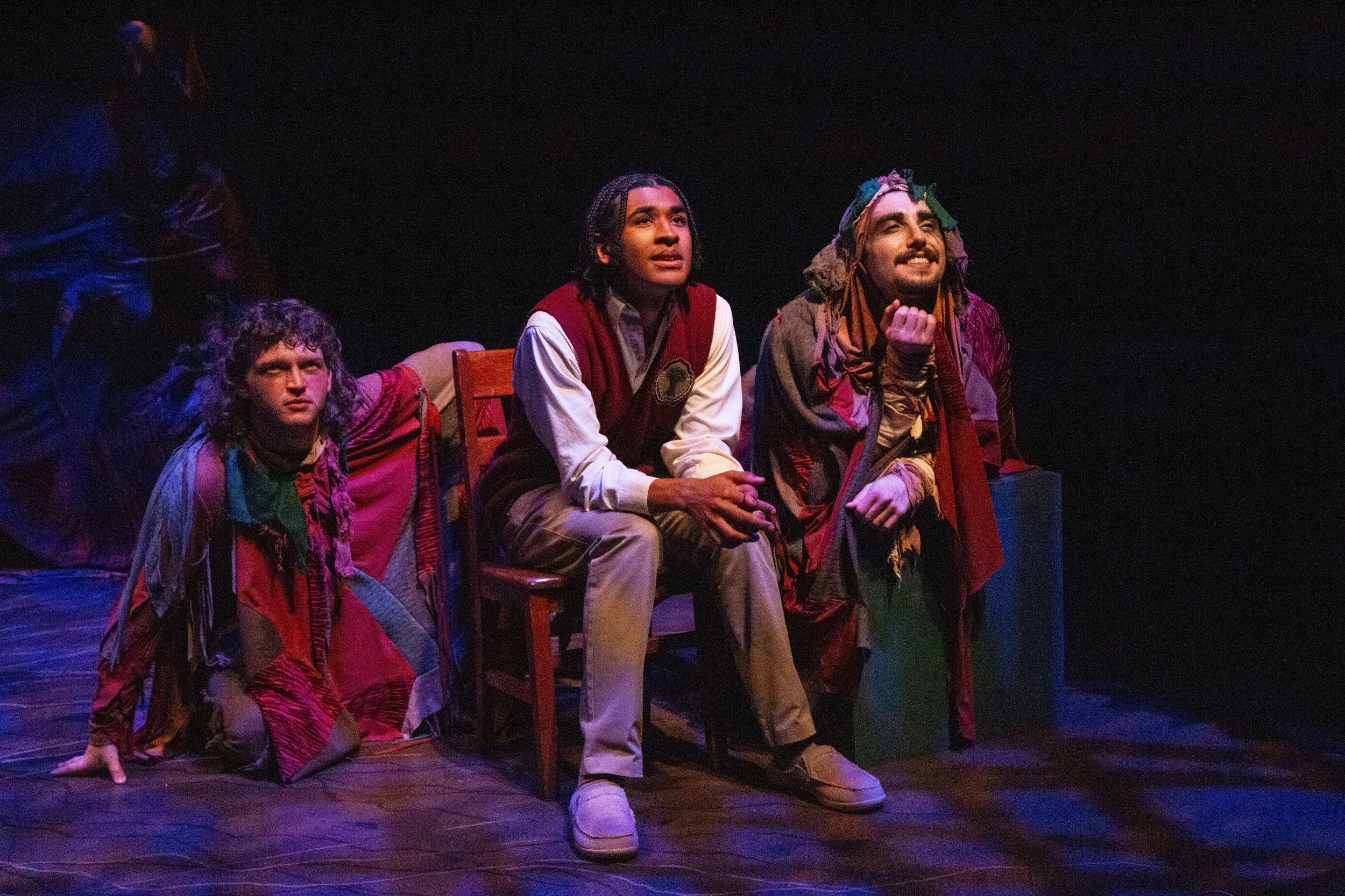 Three actors on stage in a theatrical performance, seated and kneeling, under dramatic lighting.