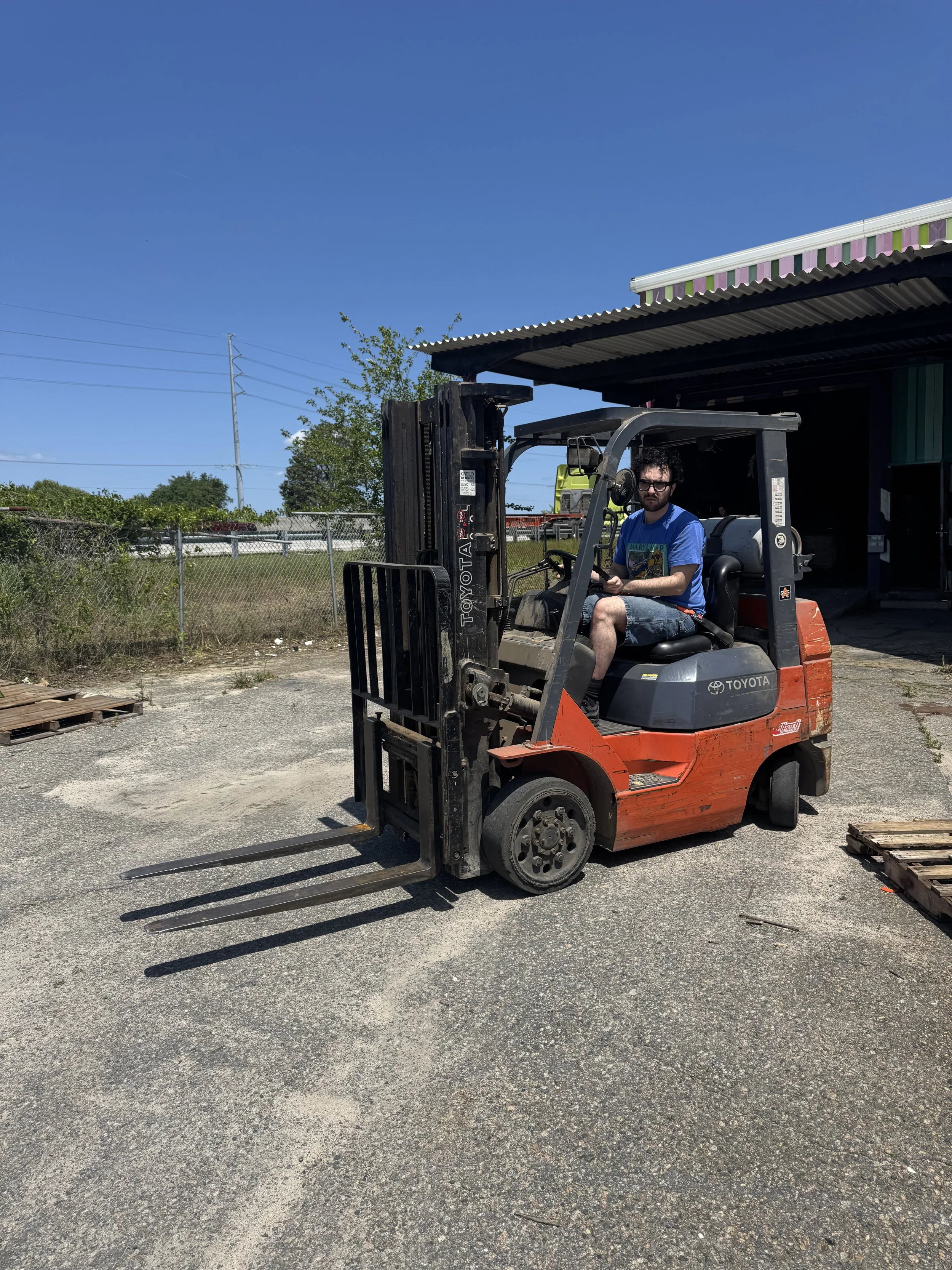 A man with dark curly hair, wearing sunglasses, a blue t-shirt, and shorts, is sitting on a red and black Toyota forklift in an outdoor industrial area under a clear blue sky.