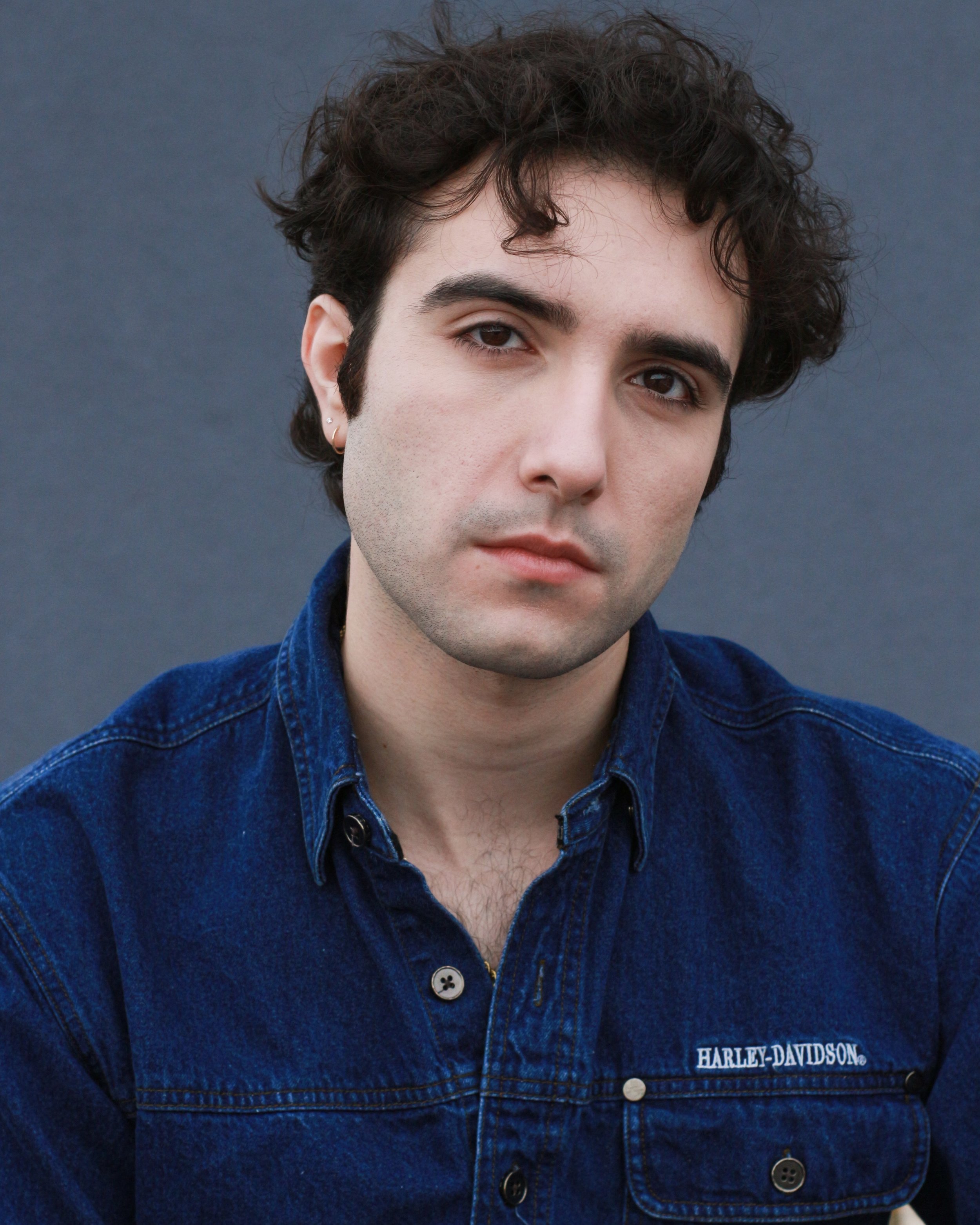 Portrait of a young man with dark, curly hair, light skin, wearing a blue denim Harley-Davidson jacket, looking at the camera with a neutral expression against a gray background.