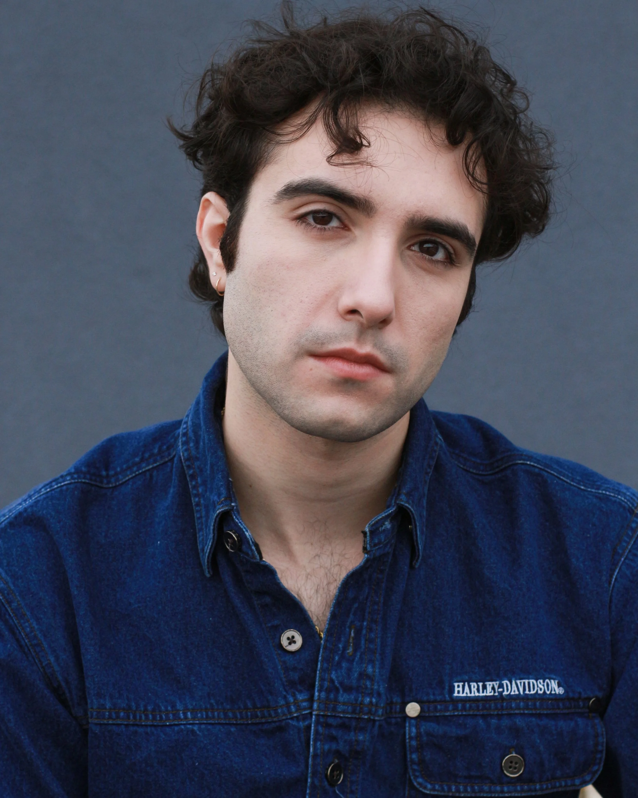 Portrait of a young man with dark curly hair, light skin, and an earring, wearing a blue denim Harley-Davidson shirt, against a gray background.