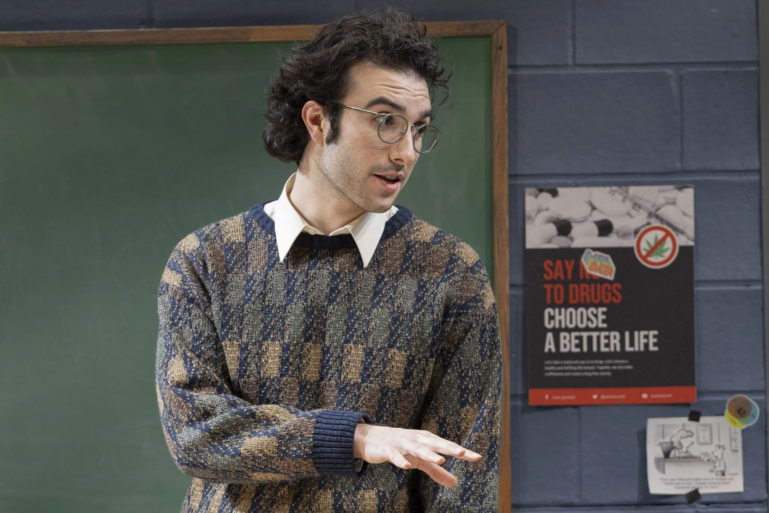 Young man with curly hair and glasses wearing a patterned sweater standing in front of a green chalkboard and a poster saying "Say No to Drugs, Choose a Better Life" in a classroom.