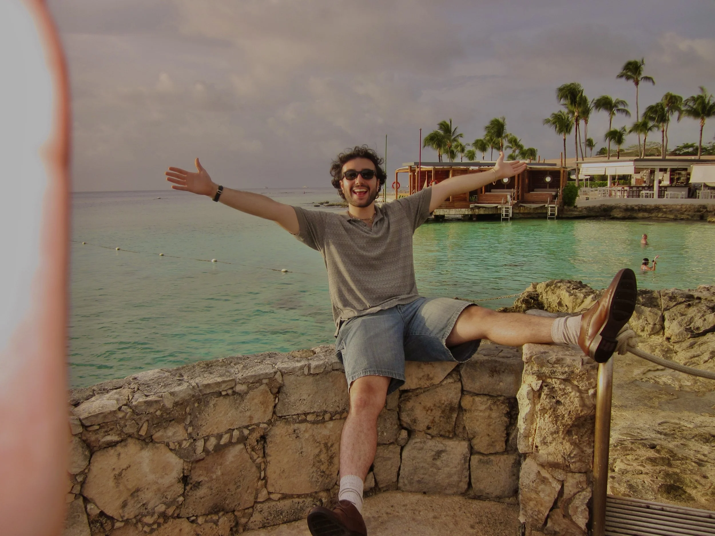 A young man with curly hair, sunglasses, a gray t-shirt, and denim shorts sitting on a stone wall by the water, with arms outstretched and smiling. Behind him are palm trees, a waterfront restaurant, and people swimming in the ocean.