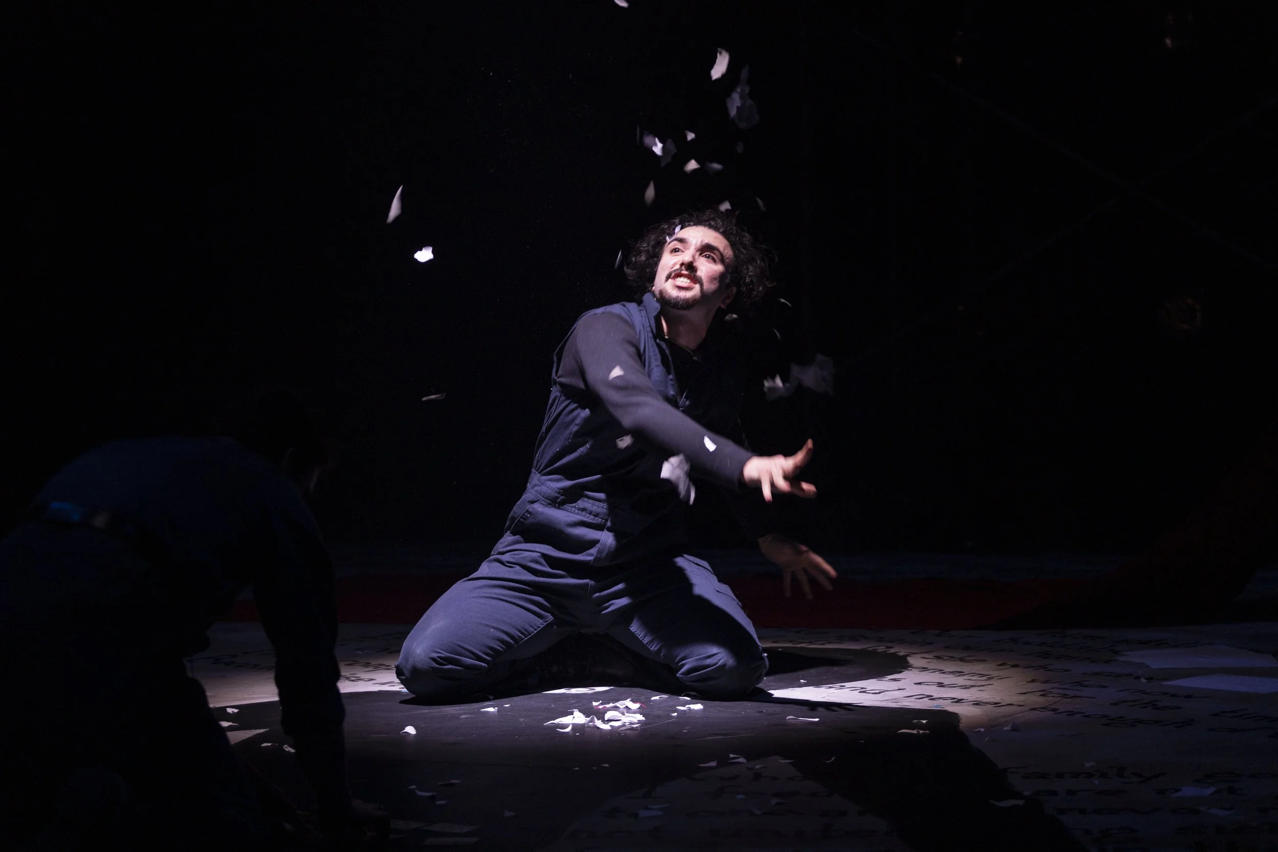 Man with dark curly hair and beard kneeling on stage, reaching out, surrounded by falling paper or confetti, dimly lit with a spotlight.
