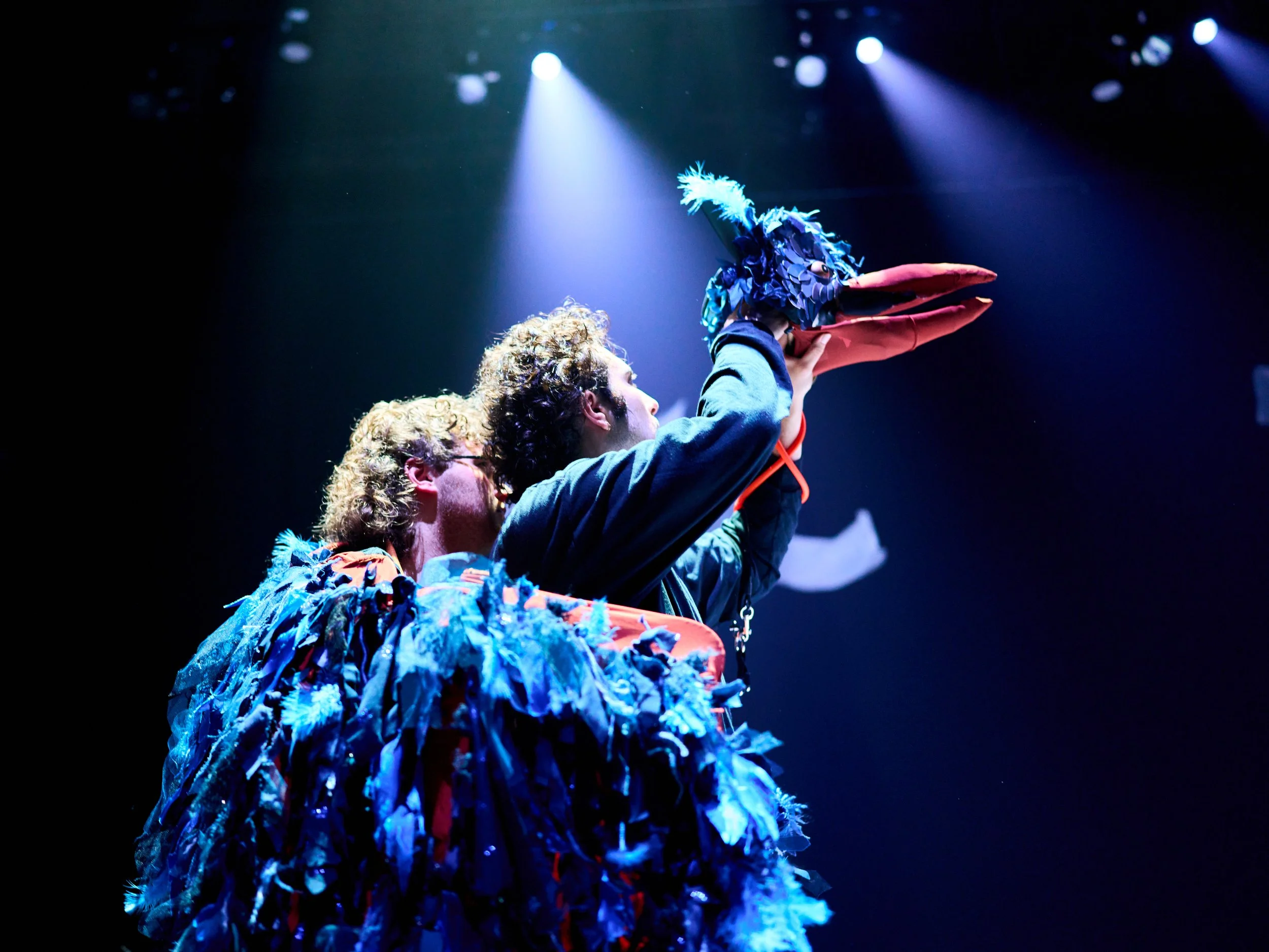 Two actors on stage, one holding a puppet resembling a bird with blue feathers, a beak, and a feathered headpiece, illuminated by stage lights.