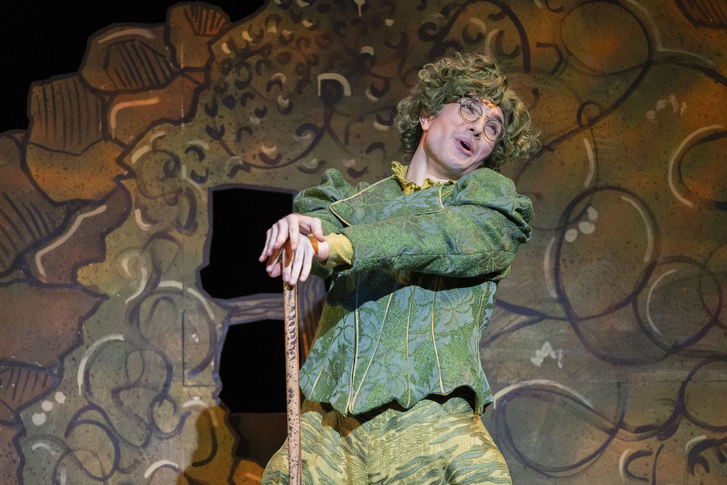 Performer dressed as a whimsical forest character with curly hair, glasses, and a flowered green outfit, performing on stage with a painted woodland backdrop.