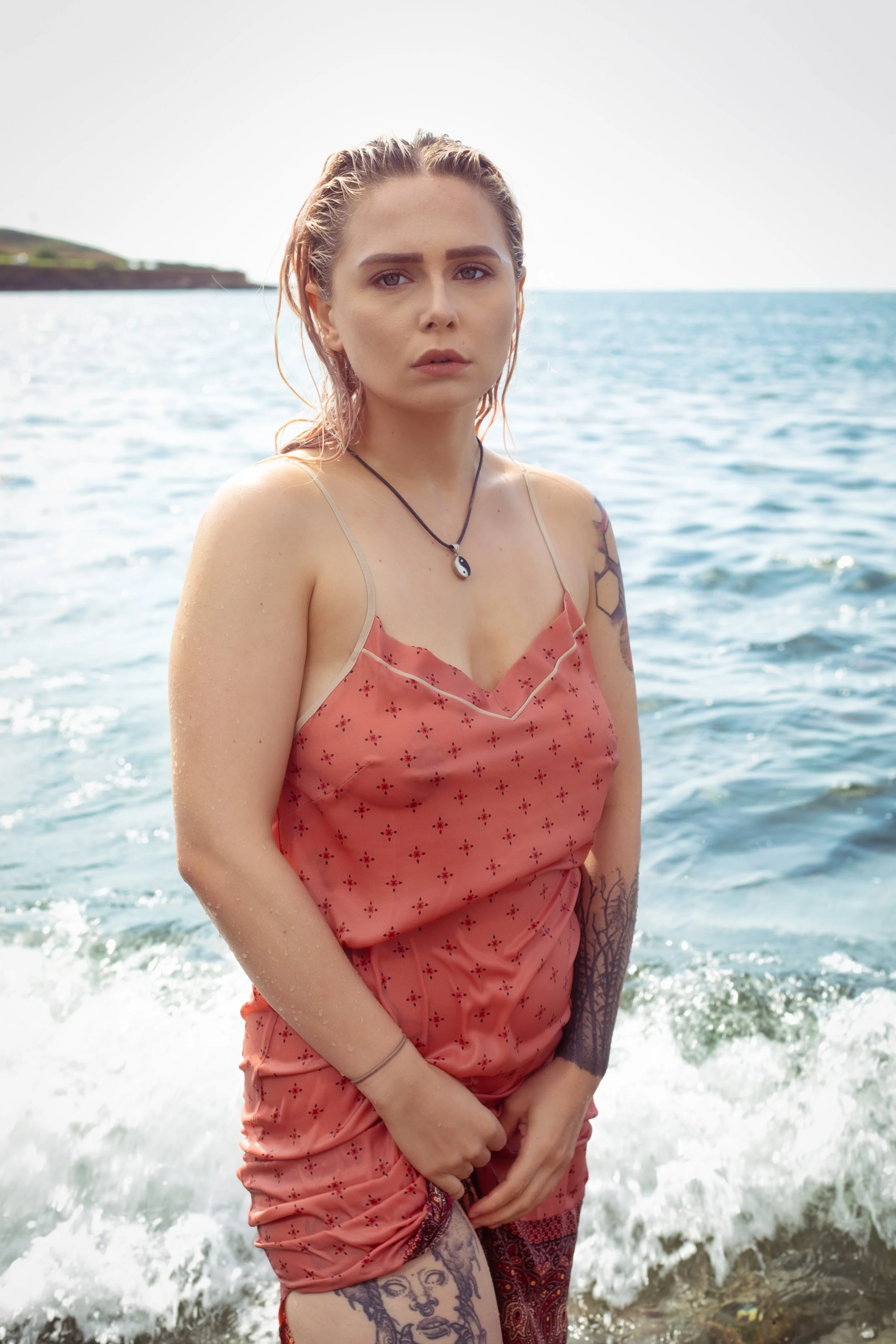 A photo of young woman with wet hair standing in the ocean near the shore, wearing a coral-colored dress with thin straps and a yin-yang necklace, with tattoos visible on her arms and legs from a night wear promotion campaign shoot