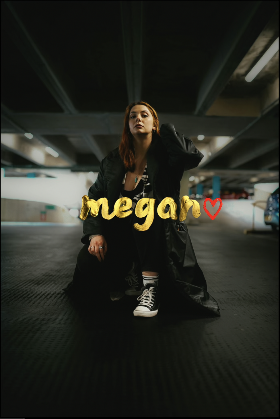 A woman in a black coat and sneakers sitting on the ground in a parking garage, with a yellow 3D text overlay that reads 'megan' and a red heart.