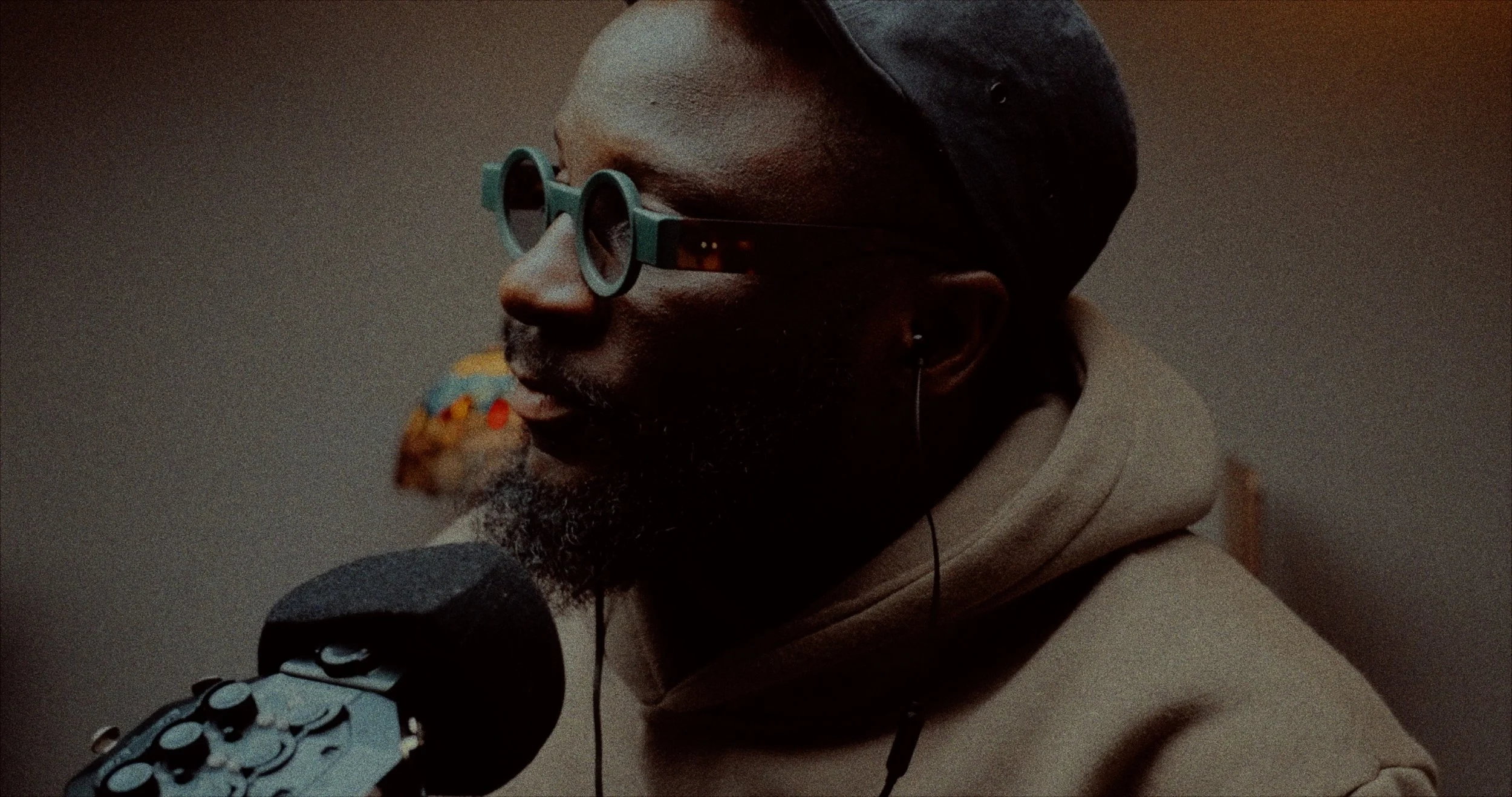 A person with dark skin, wearing gray sunglasses, earring, a head wrap, or hat, and a beige hoodie, speaking into a microphone with sound editing equipment.