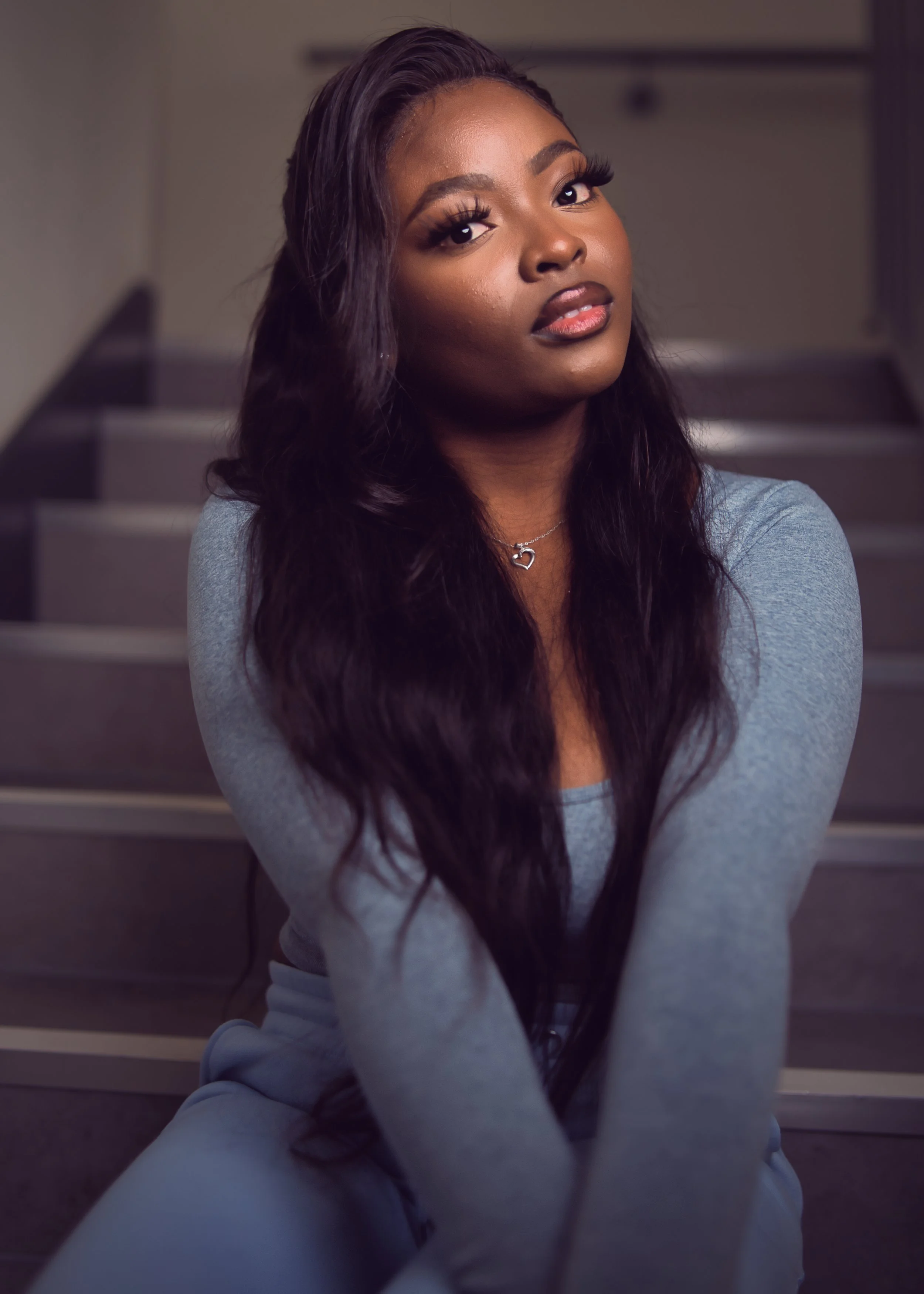 Young woman with long dark wavy hair sitting on stairs, wearing a gray long-sleeve shirt, silver necklace with a heart pendant, looking at the camera.