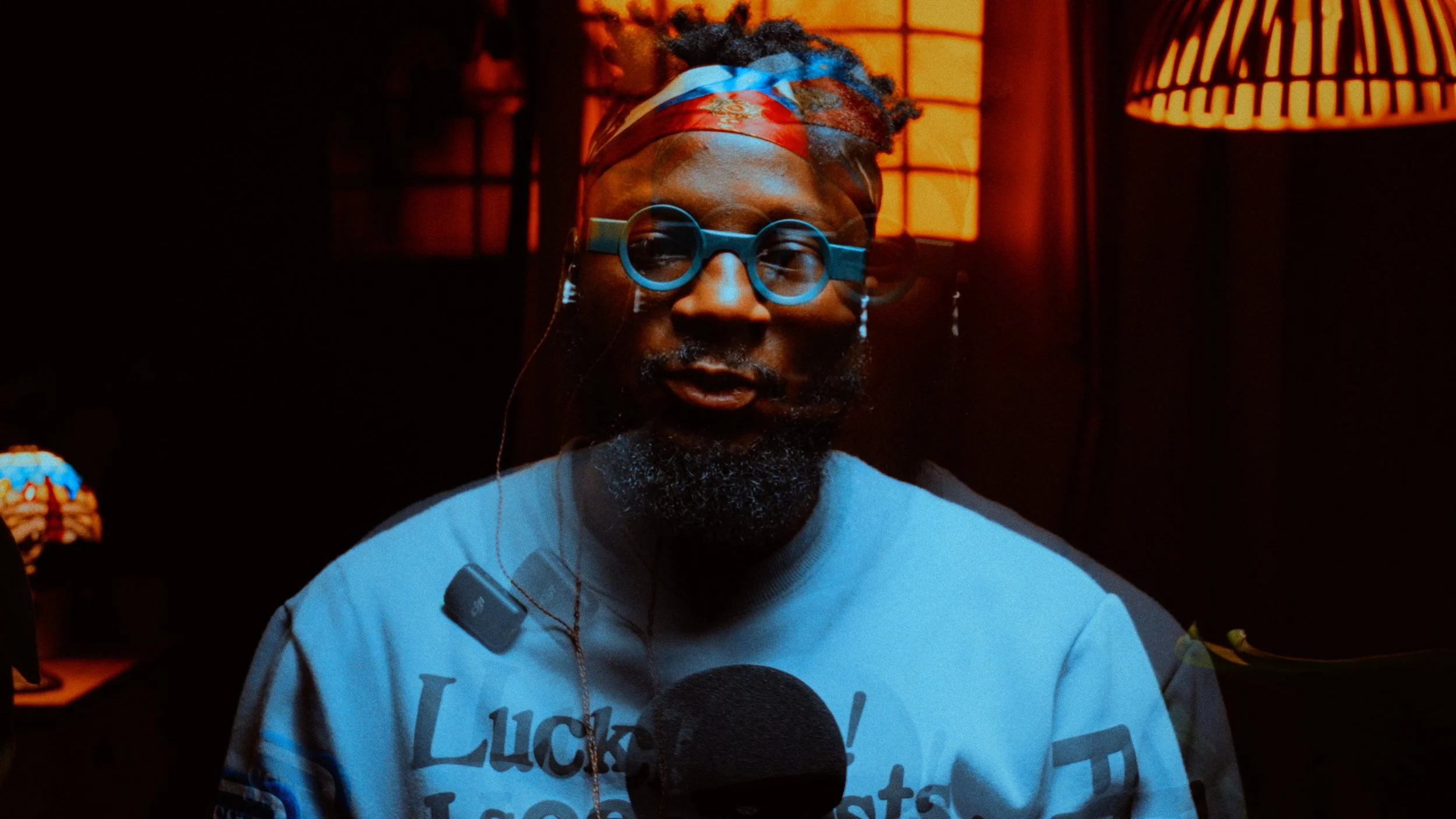 A man with dreadlocks wearing round blue glasses and a red bandana, speaking into a microphone in a dimly lit room with warm orange lighting.