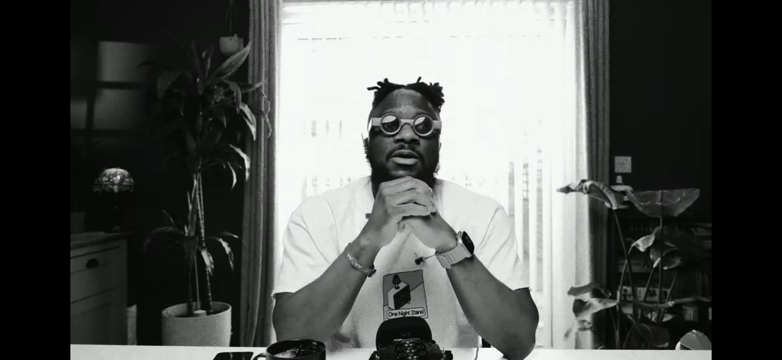 Black and white photo of a man sitting at a desk, wearing round glasses, a watch, and a T-shirt with a graphic. He has his hands clasped under his chin and is looking at the camera. There are indoor plants and curtains in the background with sunlight coming through.