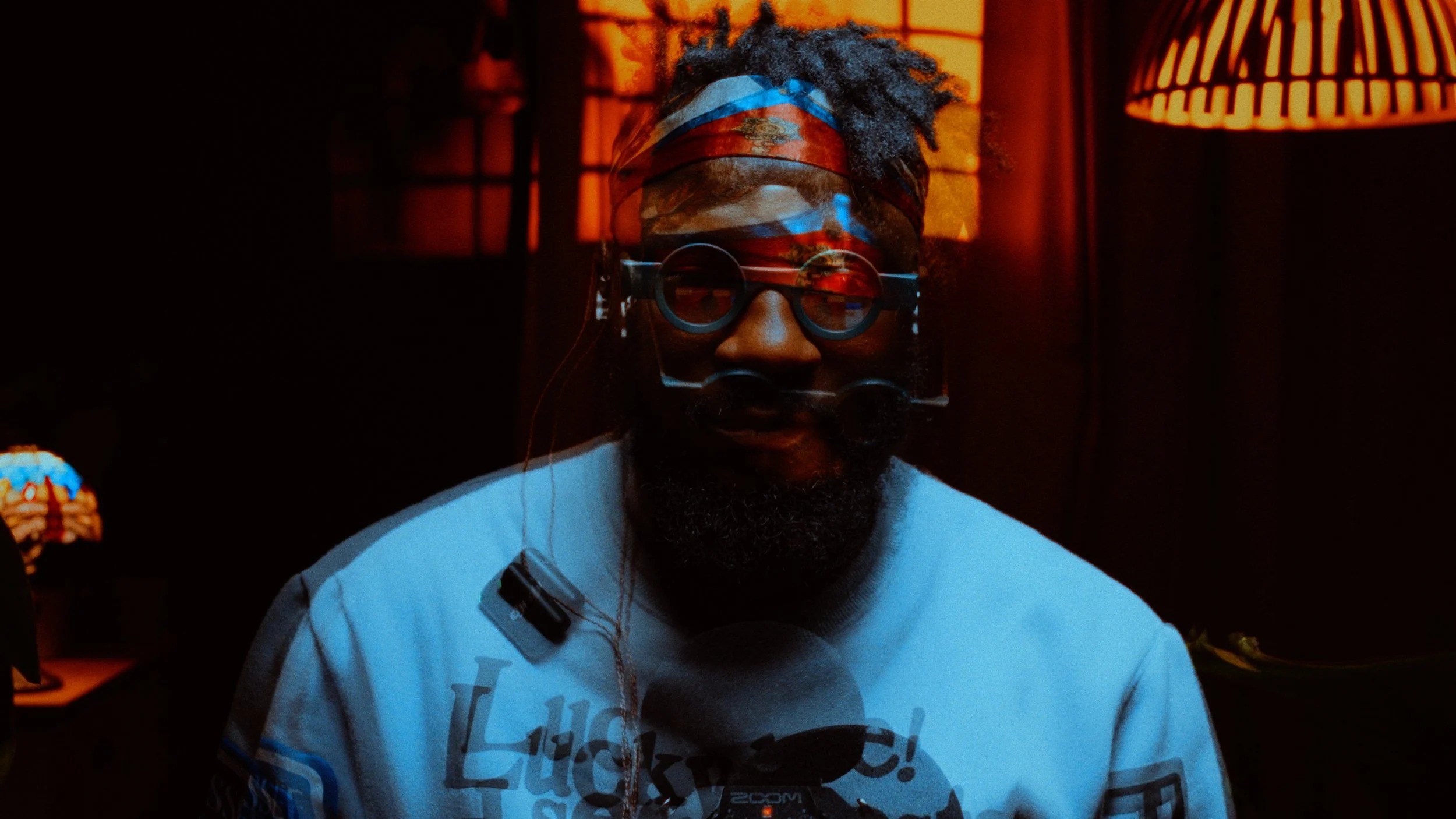 Screen grab from a talking head video of a man with a beard, wearing round glasses and a red bandana, sitting in a dimly lit room with orange and red lighting, with a microphone in front of him.