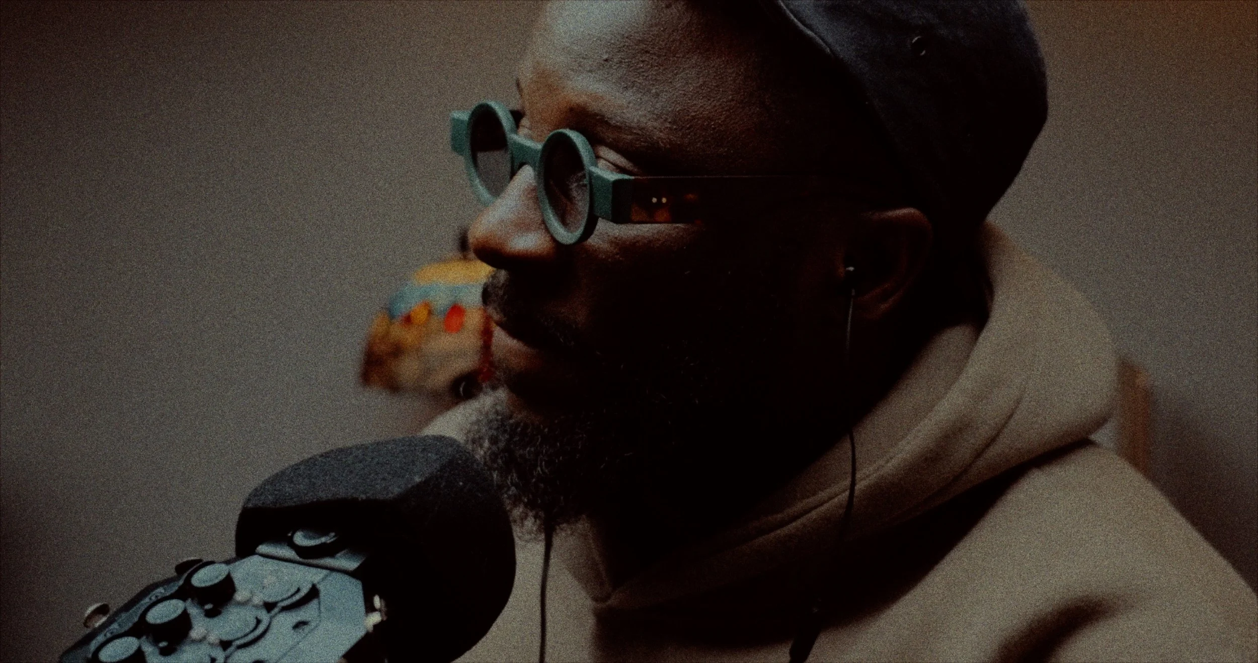 Screen grab from a talking head video of a person with dark skin wearing green sunglasses, a black headwrap, and beige hoodie, speaking into a microphone with a patterned cover, possibly recording or performing.