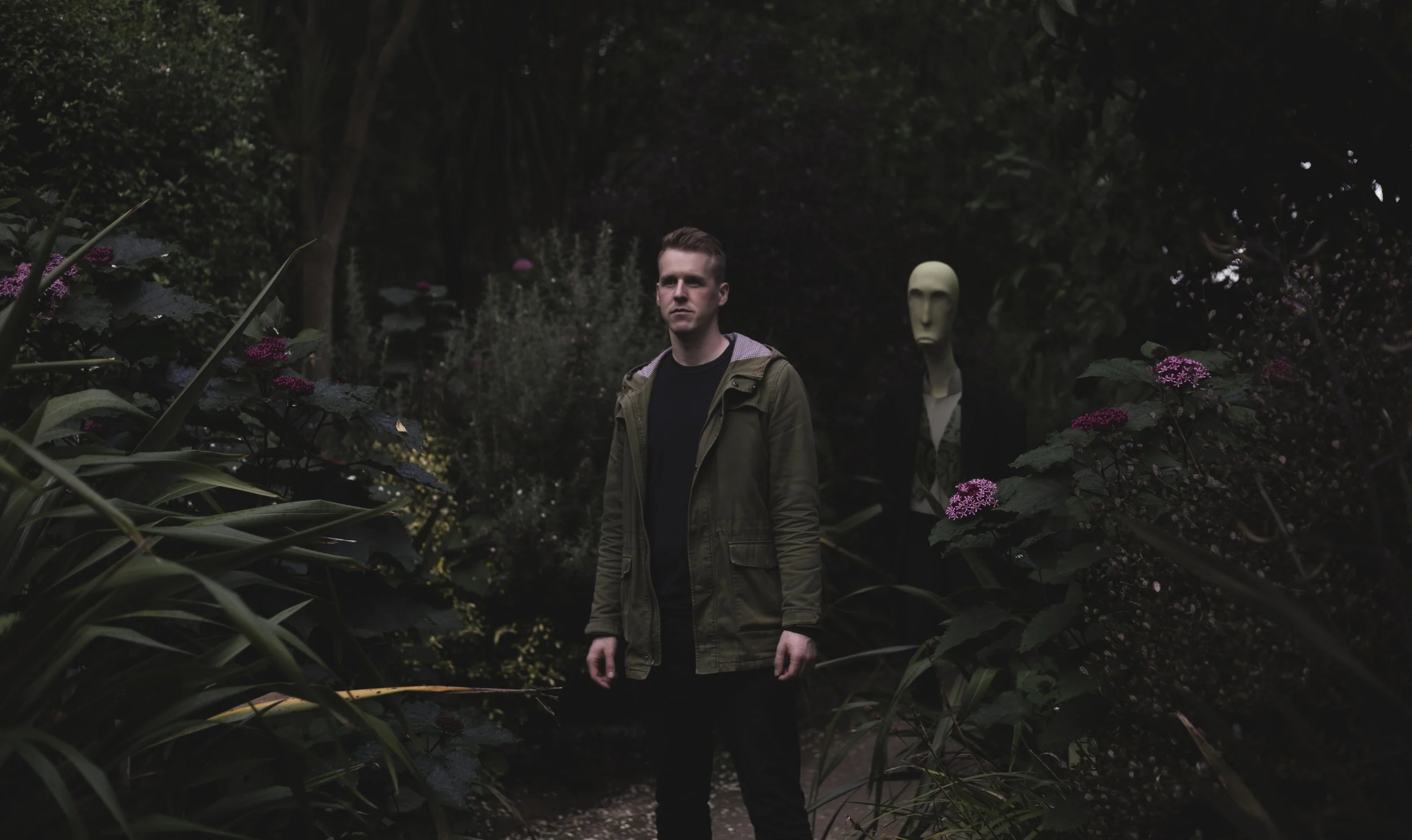 Screen grab from a music video of a man in a green jacket standing in a garden next to a mannequin with an elongated white face and dark clothing. The garden is lush with green plants and purple flowers, and the scene appears dark and mysterious.