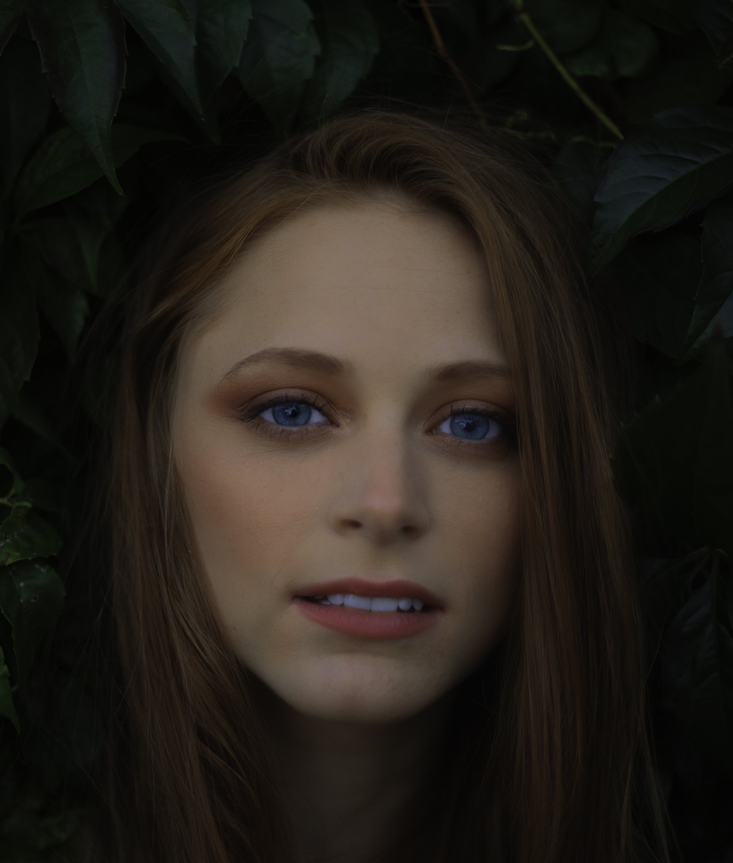 Close-up of a young woman with blue eyes and long red hair, surrounded by dark green leaves.