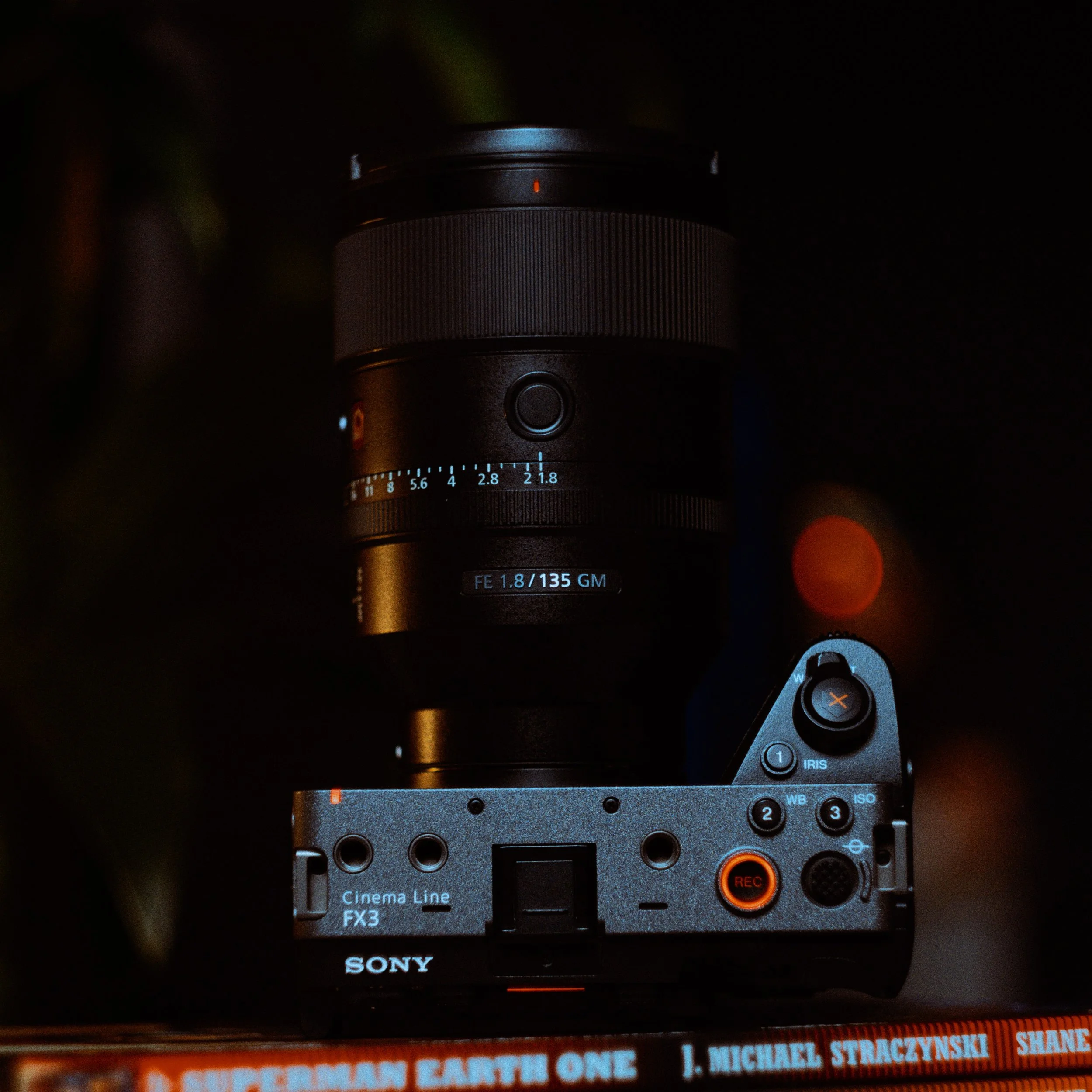 A Sony camera with a lens labeled FE 1.8/135 GM is visible, with various controls and buttons, positioned above a book titled 'Superman Earth One' by J. Michael Straczynski.