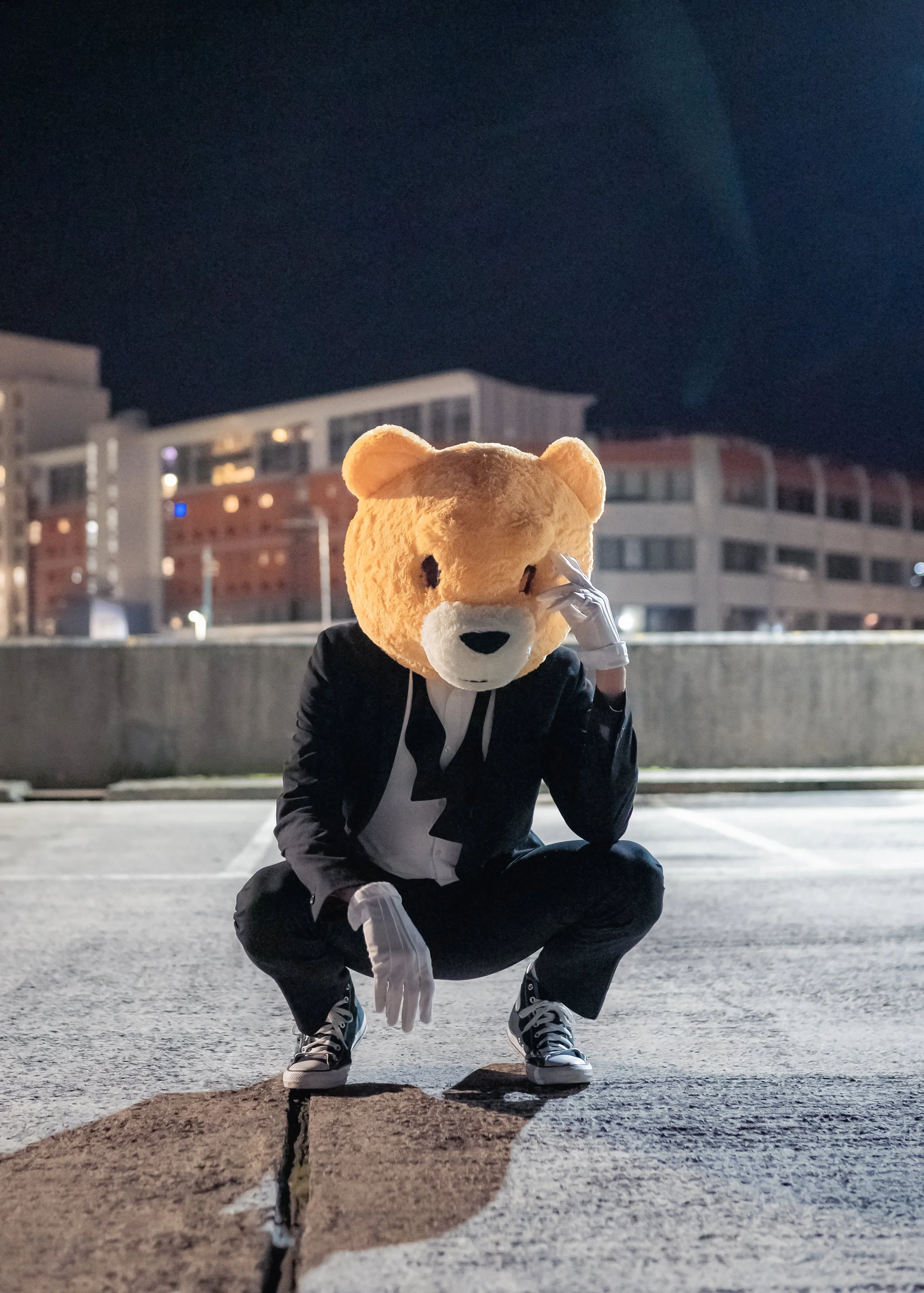 Screen grab from a music video of a person wearing a bear mask, black jacket, white gloves, and sneakers squatting outdoors at night in an urban area.