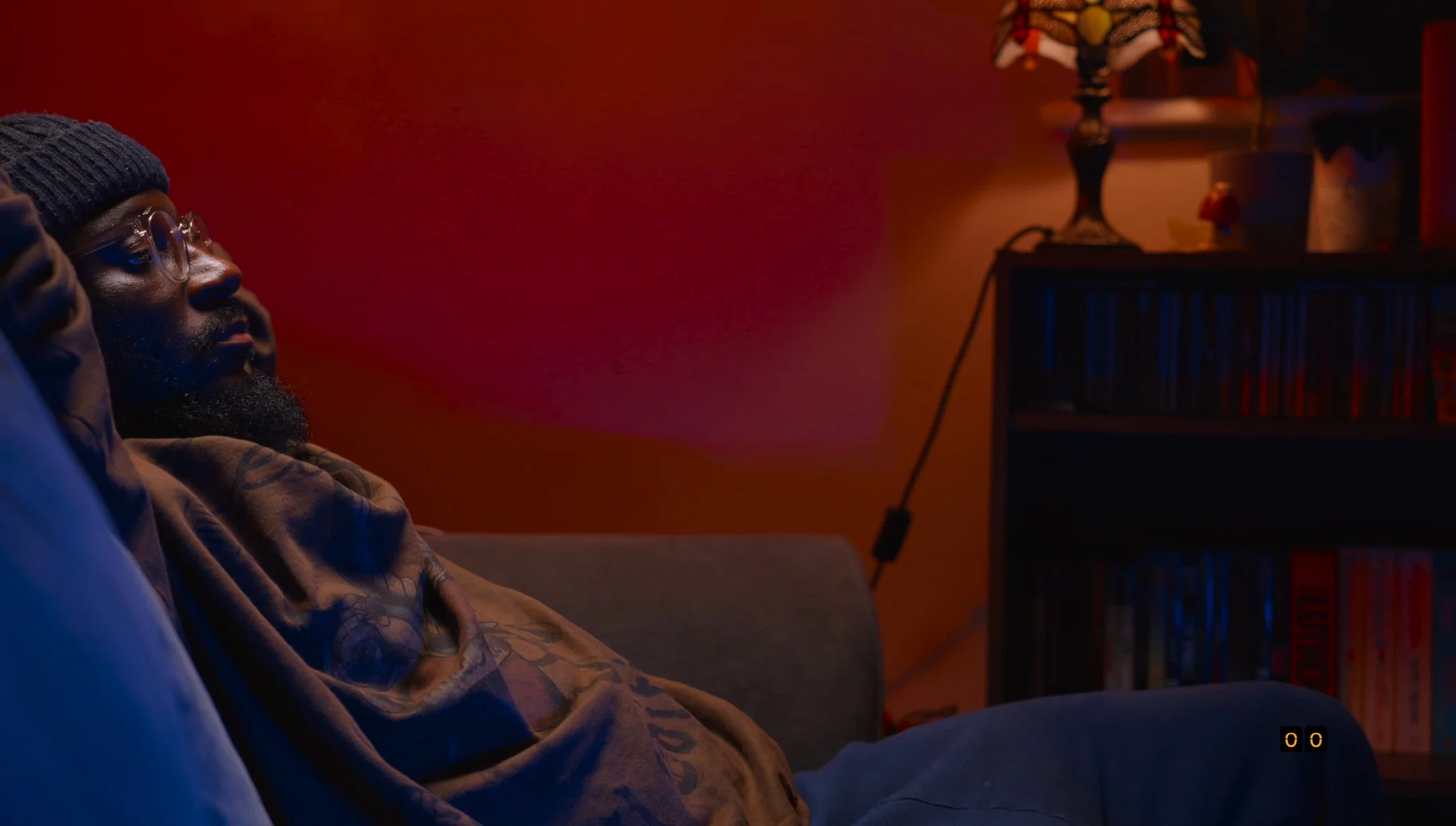 A man with glasses, a beard, and wearing a beanie is sitting on a couch in a dimly lit room, illuminated by warm, reddish lighting, with a small side table and shelves in the background.