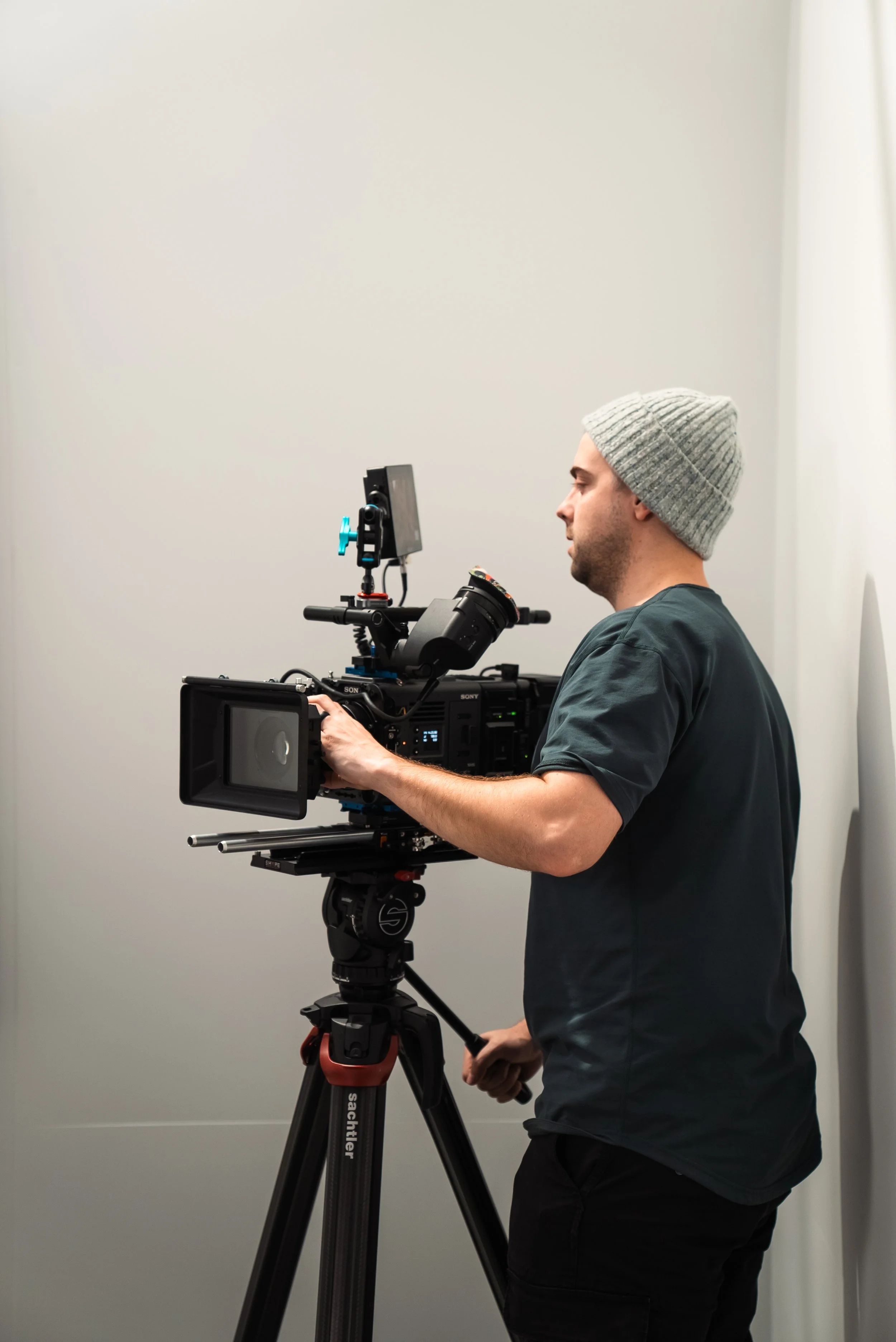 A man operating a professional video camera on a tripod in a plain room wearing a gray beanie and dark T-shirt.