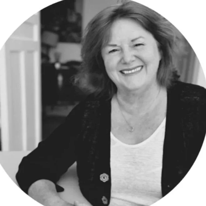 A black and white photo of a smiling woman with shoulder-length hair, wearing a light-colored top and dark cardigan, sitting at a table in a cozy room.
