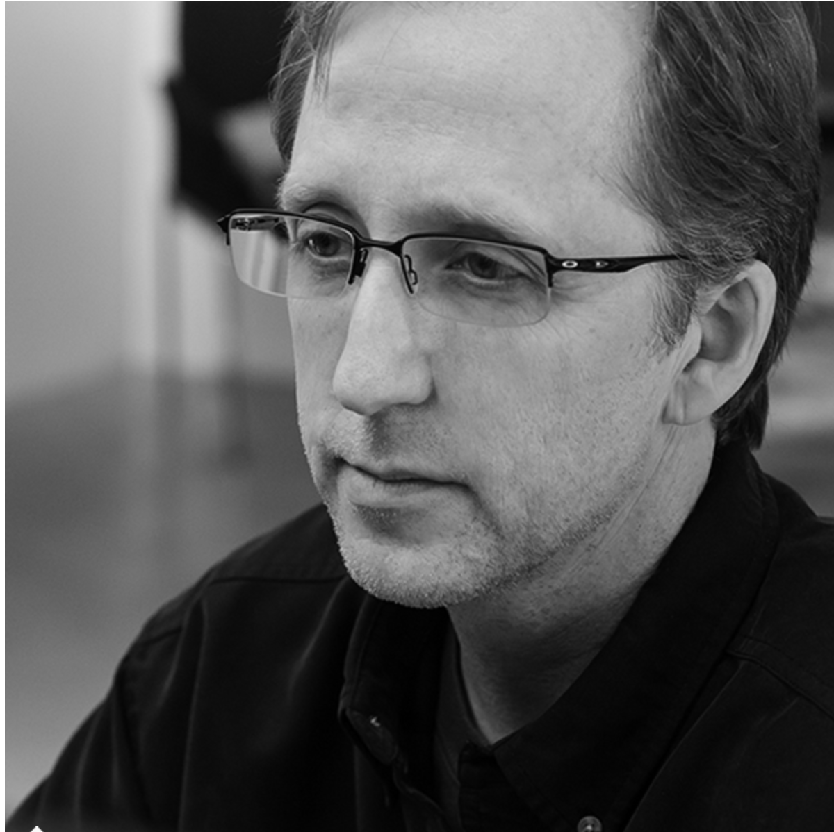 A black and white close-up of a man with glasses, short hair, and light facial hair, looking down with a thoughtful expression.