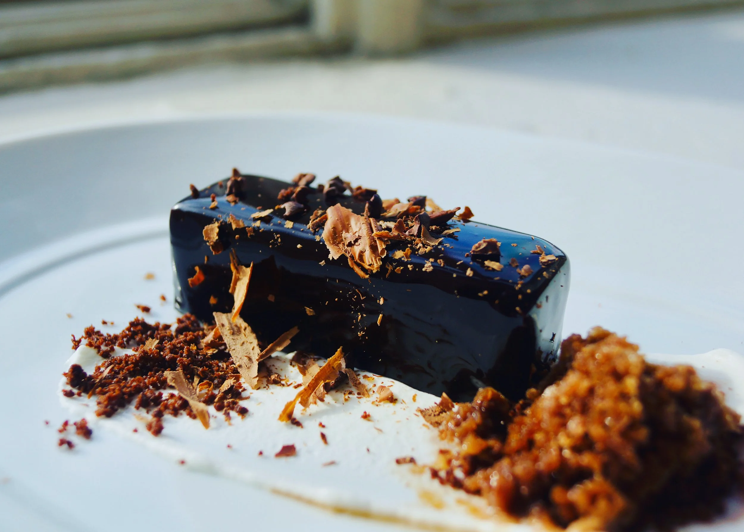 Close-up of a chocolate dessert with chocolate shavings and crumbs on a white plate.