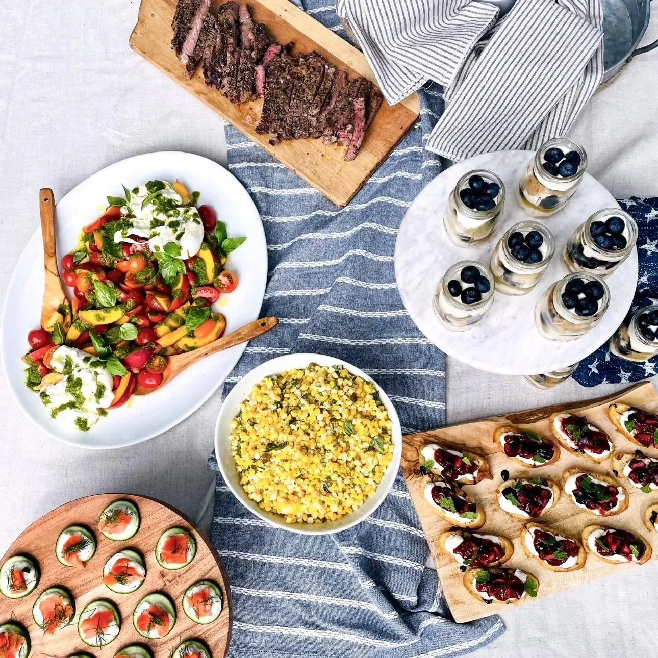 A table with a variety of foods including sliced steak on a wooden board, a bowl of salad with cherry tomatoes, basil, and cheese, a bowl of corn, small glass jars with blueberries and cream, cucumber bites with smoked salmon and dill, and canapés wi