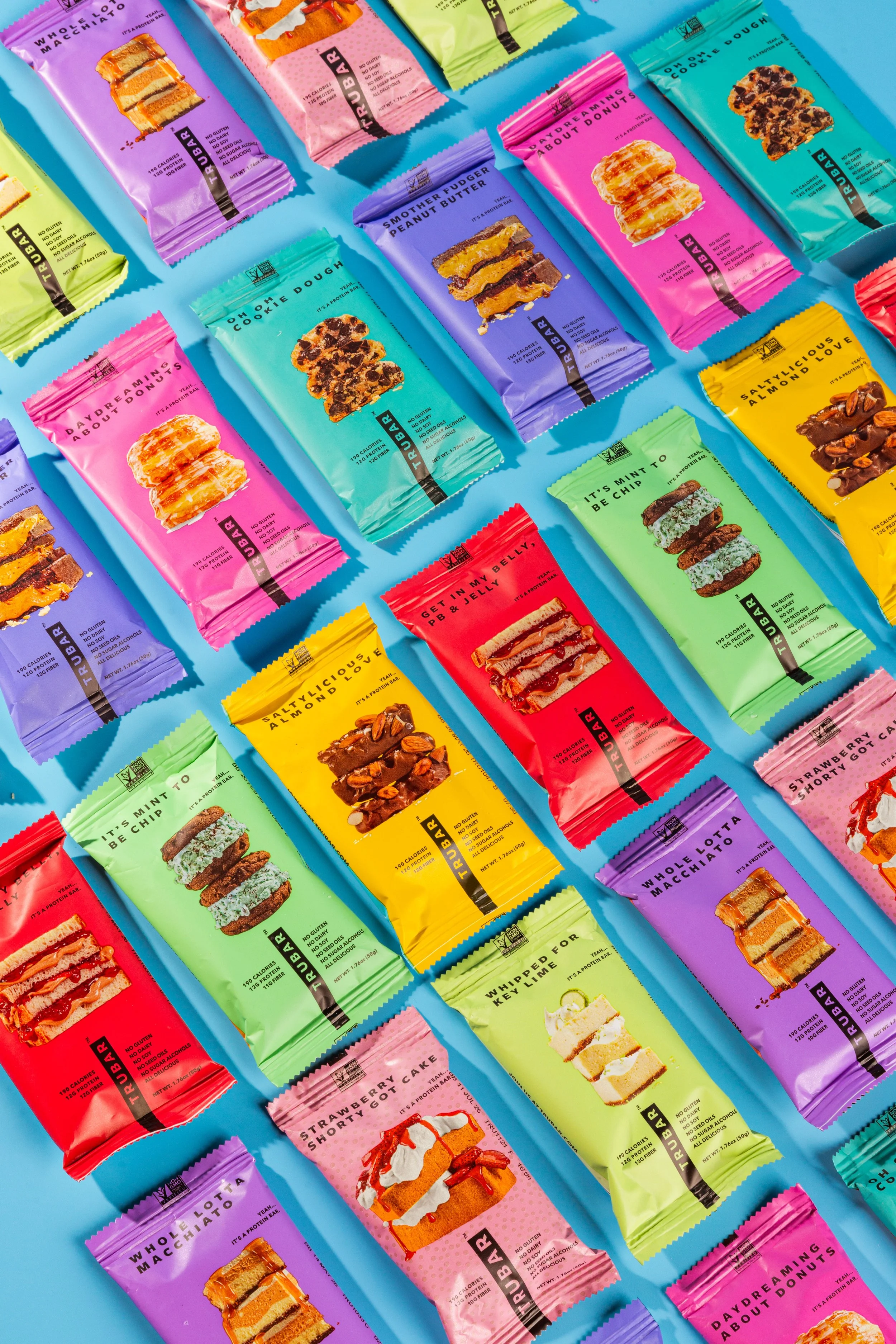 Arrangement of colorful snack bars on a blue background, with wrappers of various flavors and designs including pink, purple, green, yellow, red, and blue, each featuring images of the snack inside.