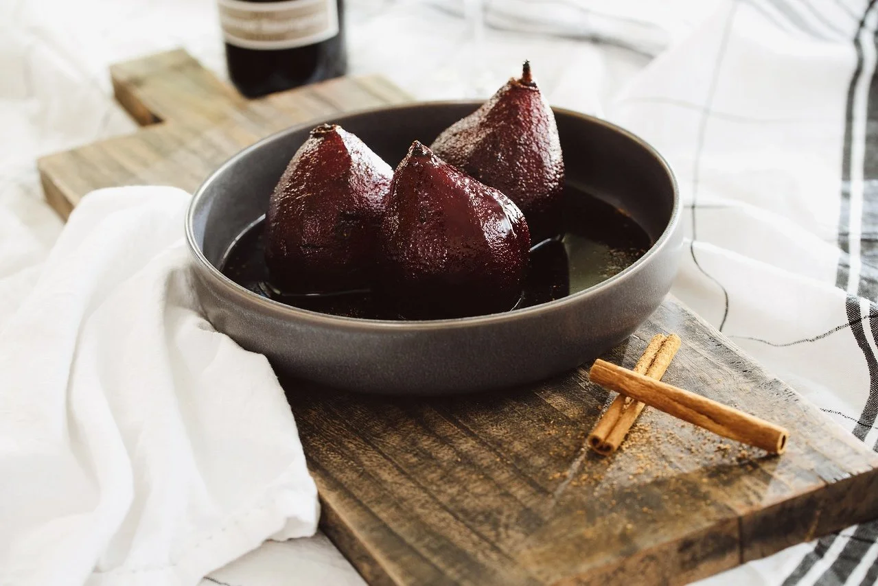 A bowl of poached pears in red wine on a wooden cutting board with cinnamon sticks.