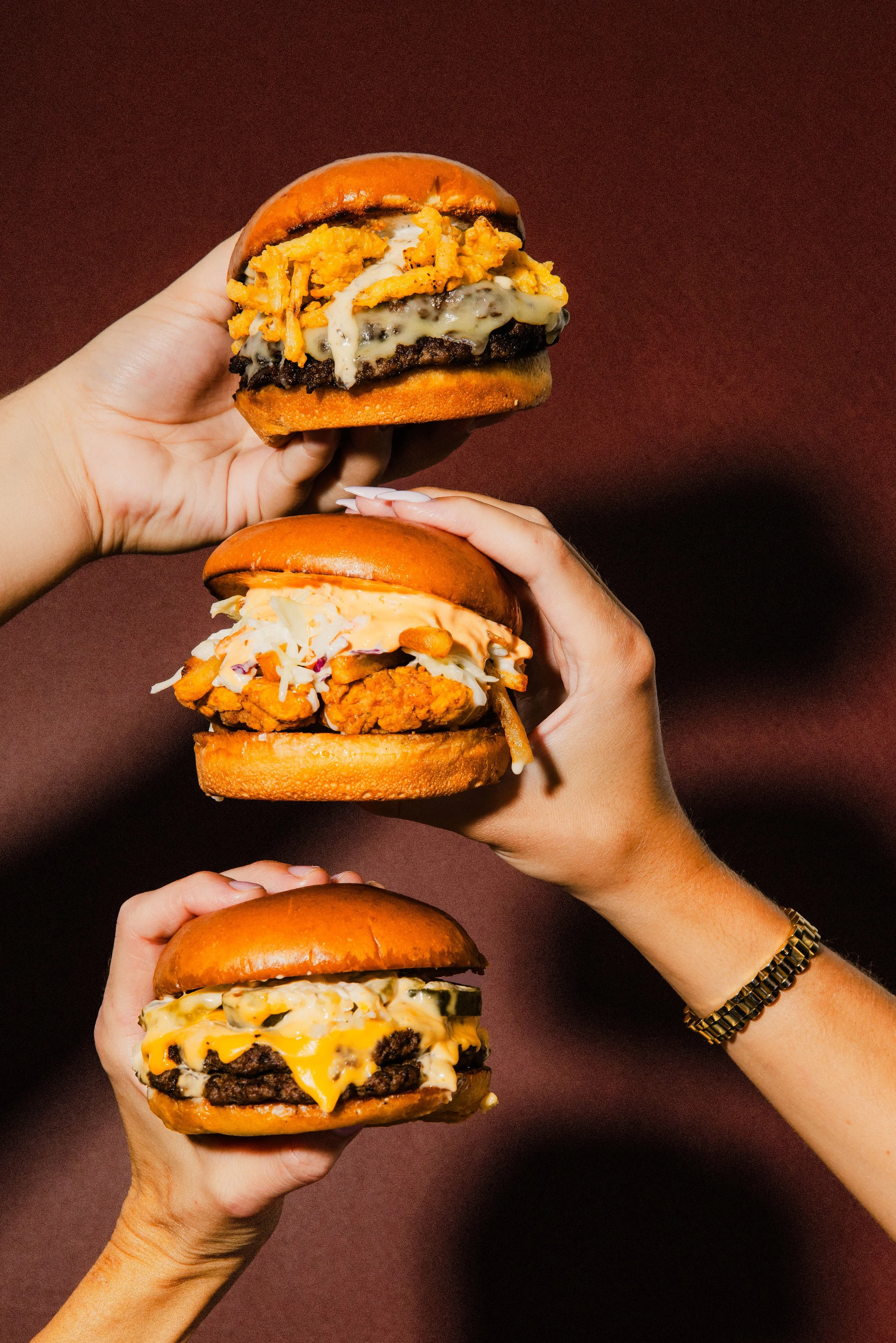 Multiple hands holding different cheeseburgers with various toppings against a dark background.