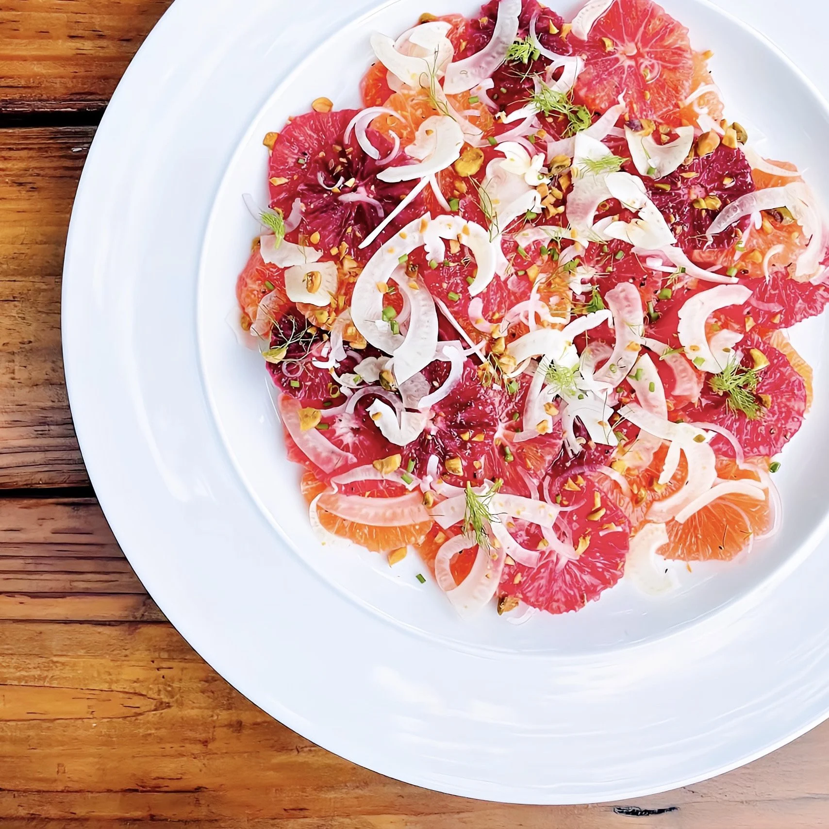 A white plate with a citrus salad containing slices of pink grapefruits, oranges, and blood oranges, topped with sliced onions, chopped nuts, and garnished with herbs, on a wooden table.